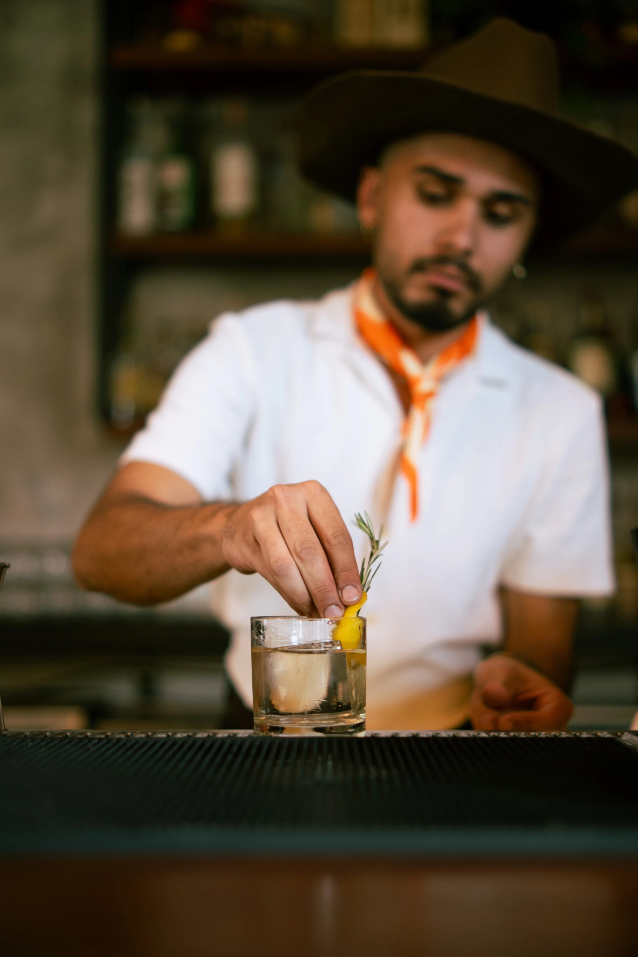 A man with a hat and orange scarf is garnishing a drink with rosemary.