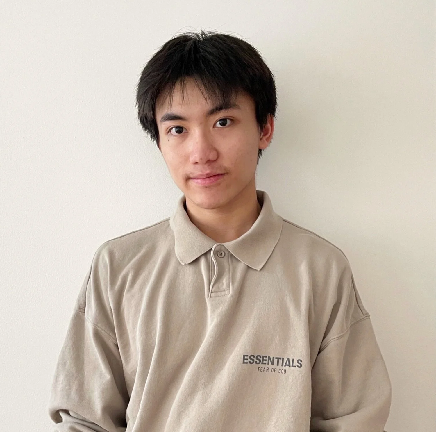 Young man with short black hair and light skin wearing a beige polo shirt with the words 'ESSENTIALS FEAR OF GOD' embroidered on the chest, standing against a plain white wall.