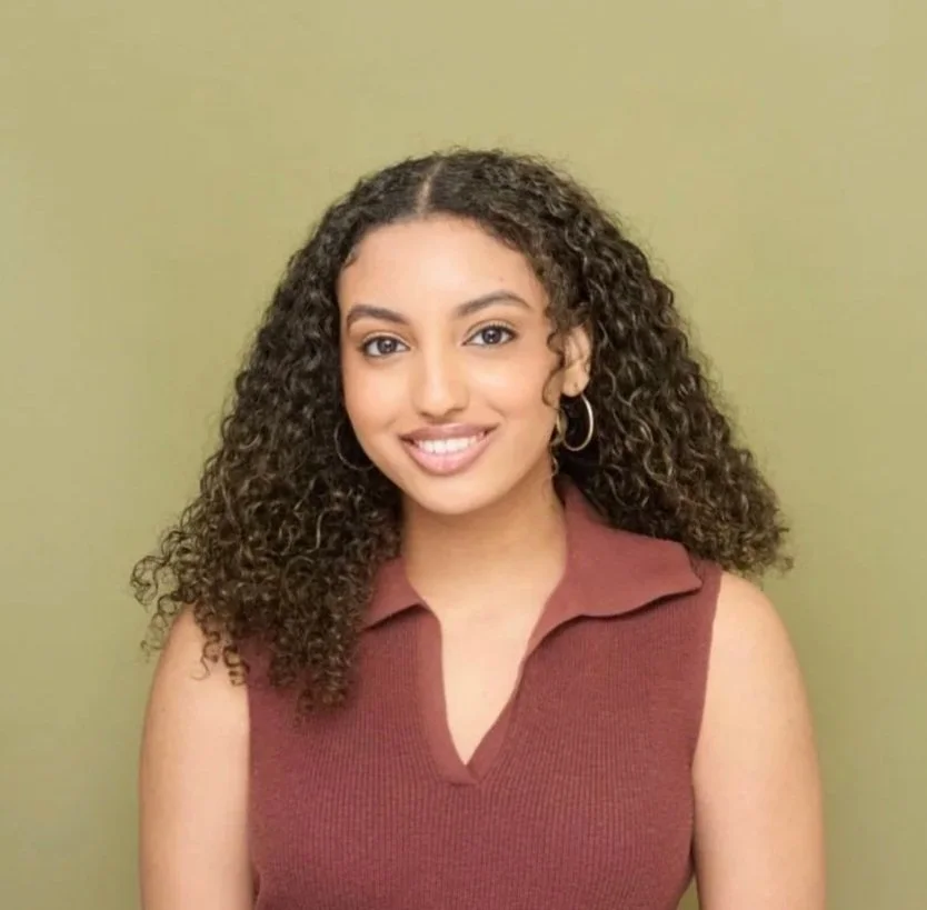Young woman with curly hair wearing a sleeveless maroon top, smiling, standing against a green background.