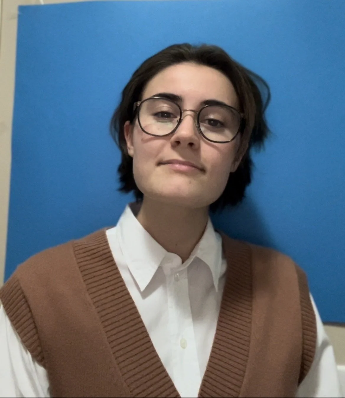 A young person with short dark hair and glasses, wearing a white collared shirt and a brown vest, standing in front of a blue background.
