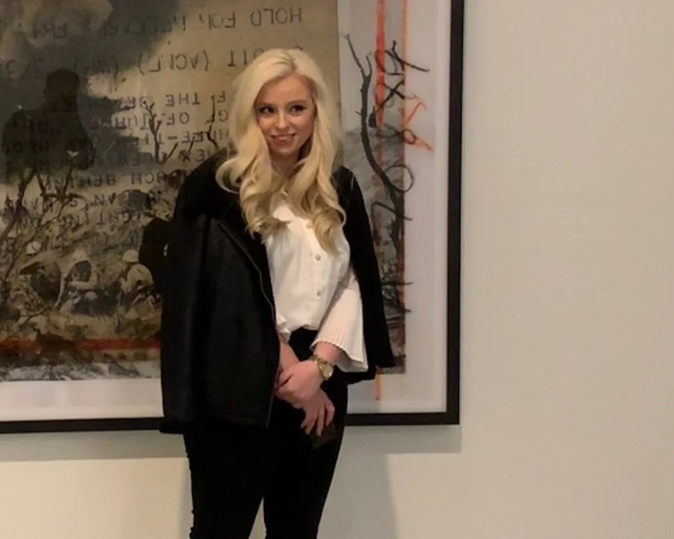 A woman with long blonde hair wearing a black jacket and white blouse standing in front of a framed artwork on a white wall.
