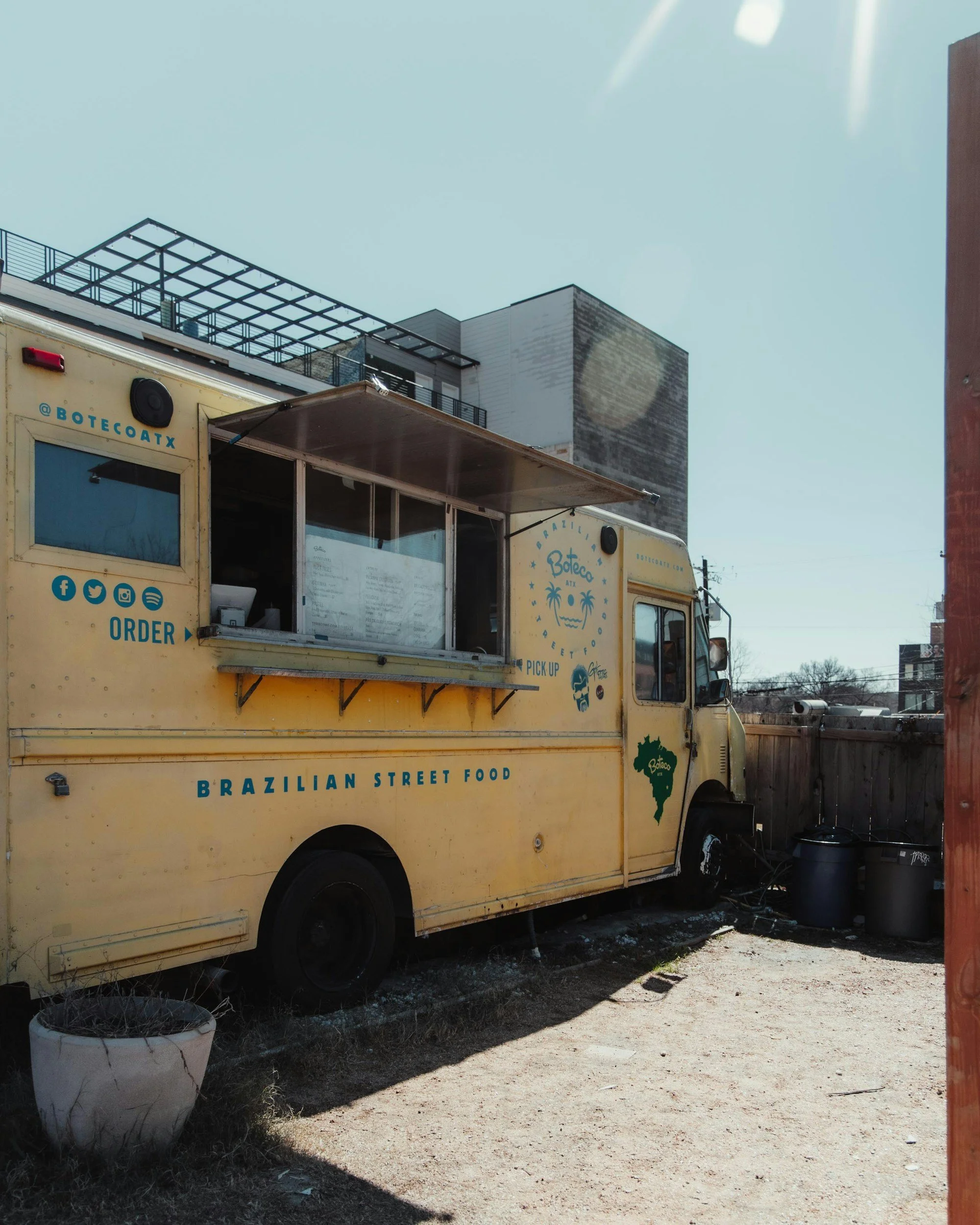 Yellow Brazilian street food food truck with open service window, instructions for ordering, and social media icons, parked on a dirt lot with a nearby fence and barrels, against a background of buildings and clear sky.