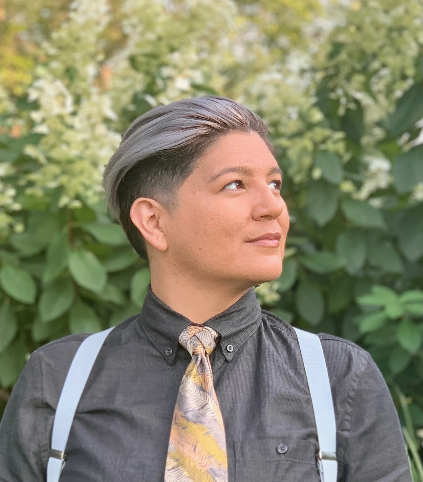 Person with short, styled gray and dark hair, wearing a dark button-up shirt with suspenders and a patterned tie, looking to the side, outdoors with green foliage in the background.
