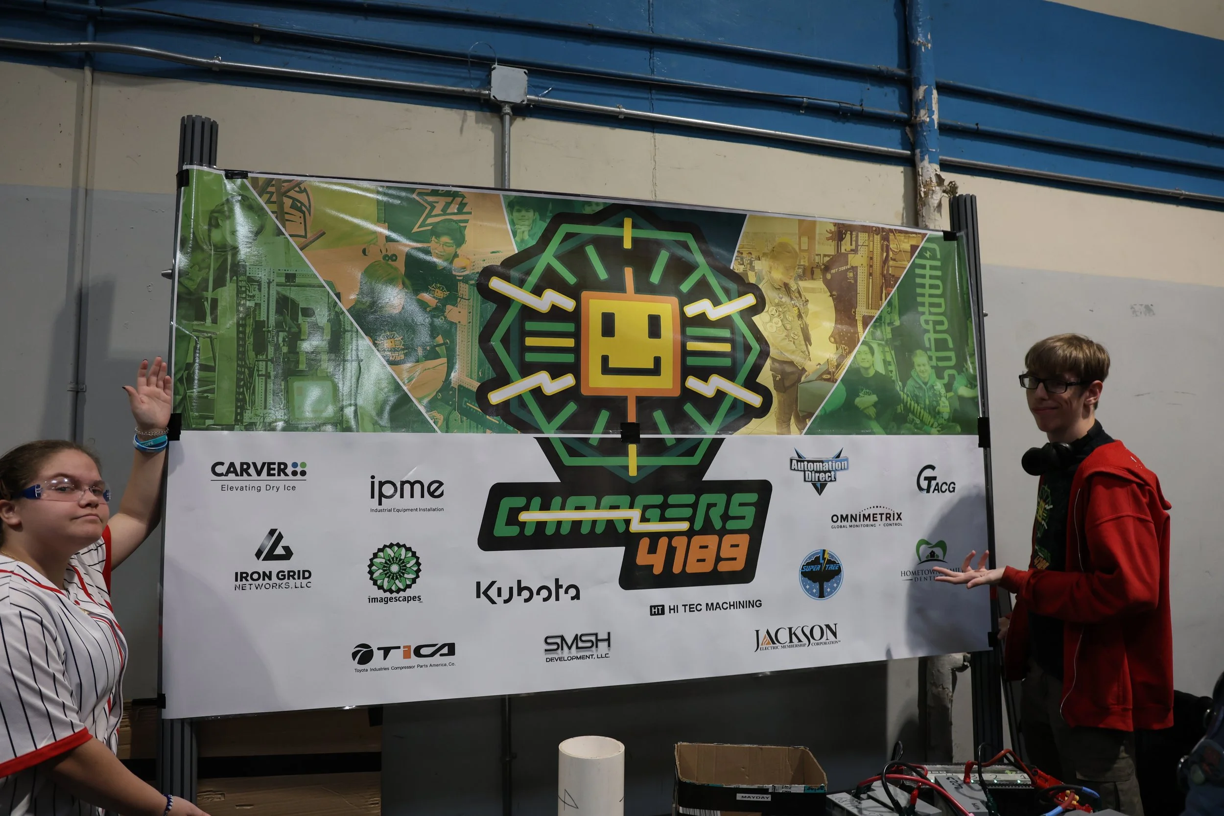 Two students standing on either side of a banner with the CyberChargers 4189 logo, surrounded by sponsor logos, at a robotics competition.