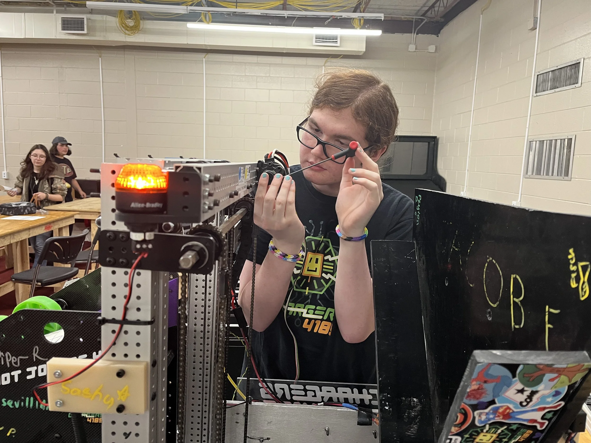 Teenager working on a robotics project at a competition, using a screwdriver to adjust a robot with a flashing orange light.