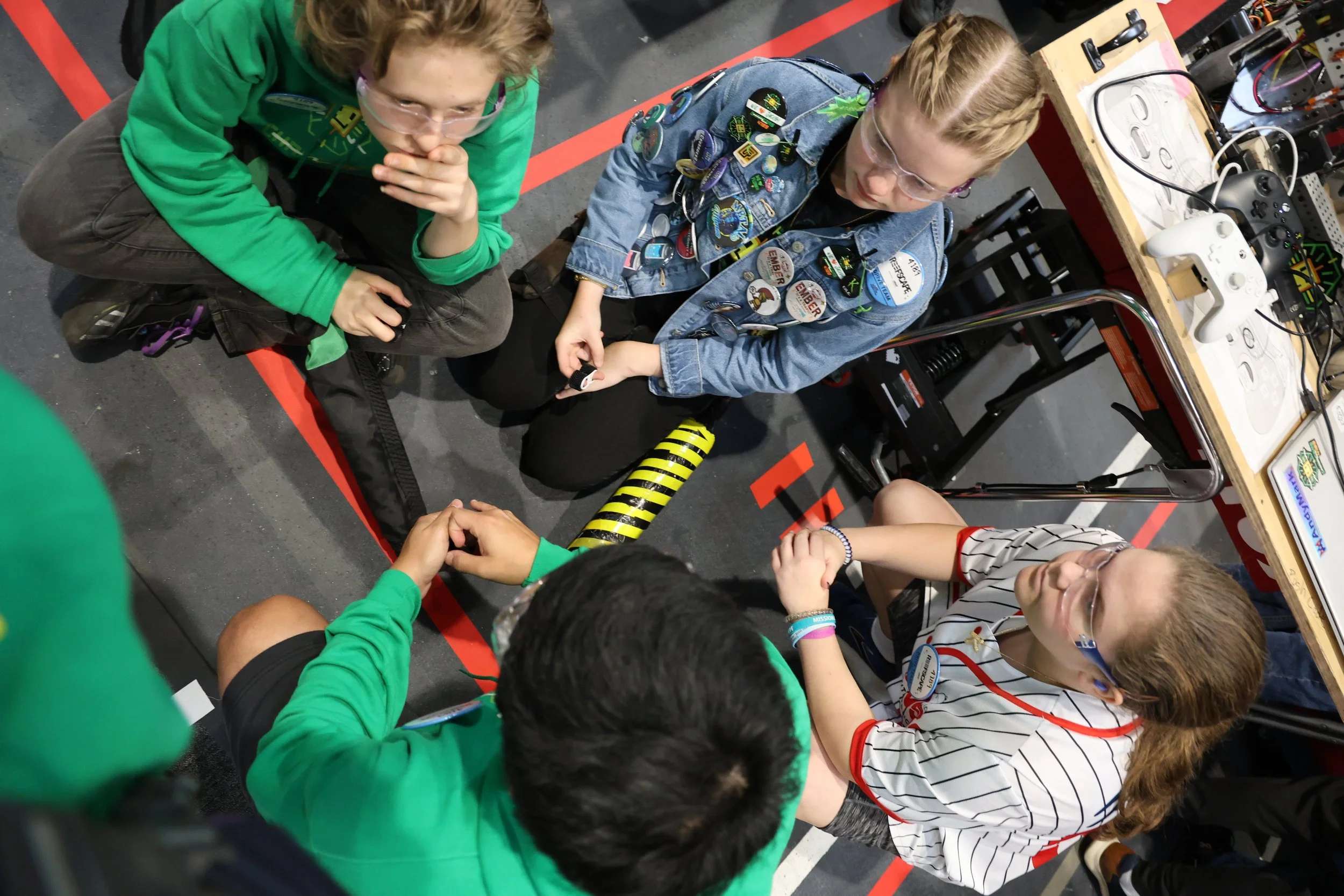 Four children sit on a gym floor in a circle, holding hands, engaged in a group activity. The children wear casual clothing, glasses, and some have wristbands. They are surrounded by robotics equipment and controllers on a nearby table.