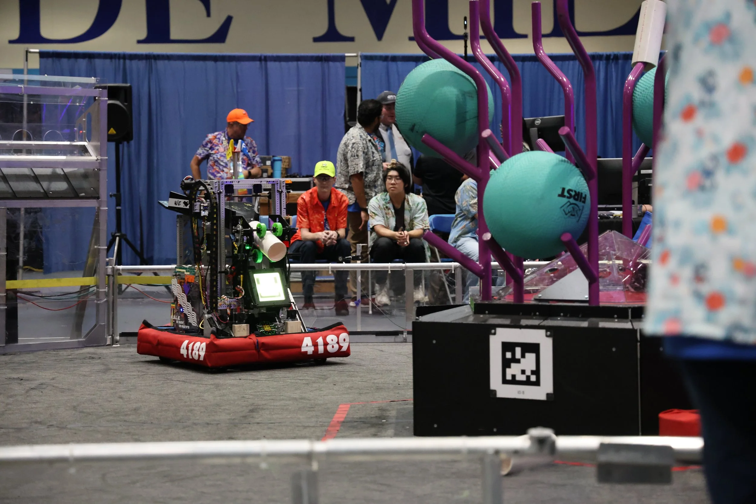 A robot with the number 4189 on a red base in a robotics competition, facing green balls on a purple rack. Several people are watching and standing behind a blue curtain, with some seated and others standing in the background.