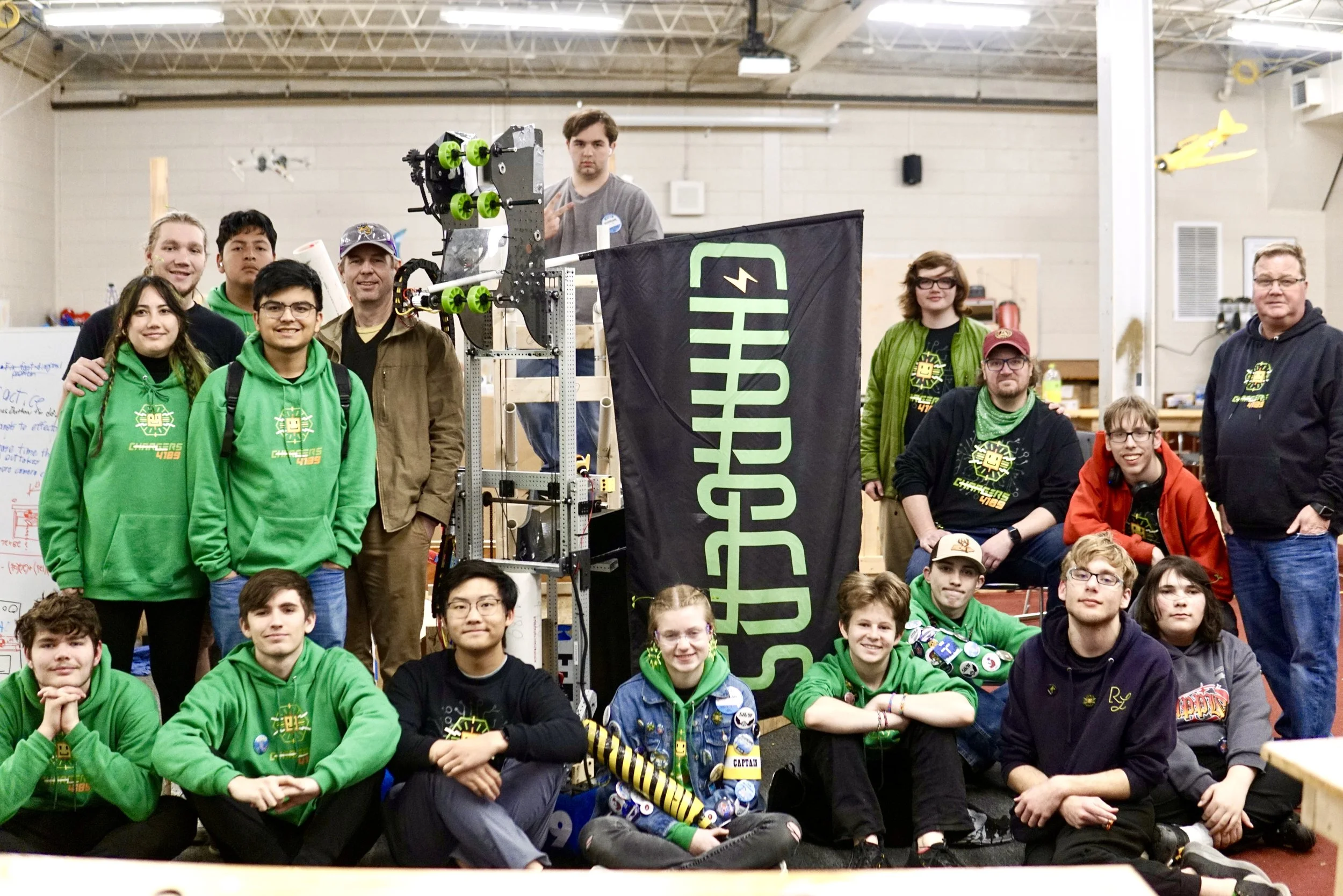 Group of young students and coaches posing with their robotics team project at a workshop, with a robot and a Challengers banner.