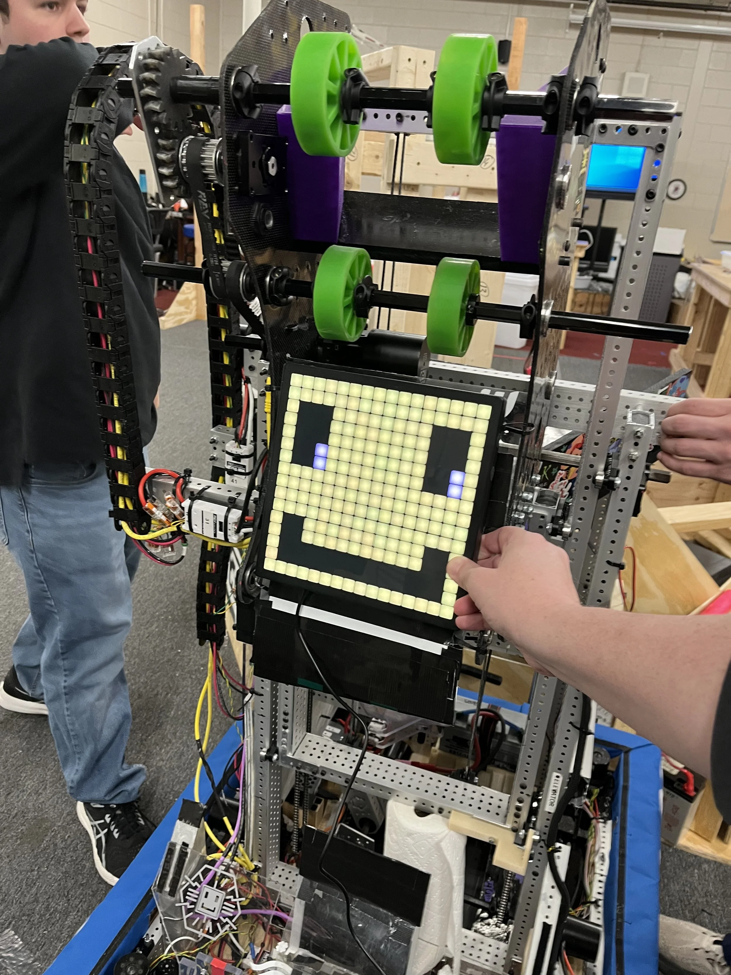 A robotics project featuring a rectangular frame with a digital pixel display showing a smiling face, green wheels, and mechanical components, with people working on it in an indoor workshop.
