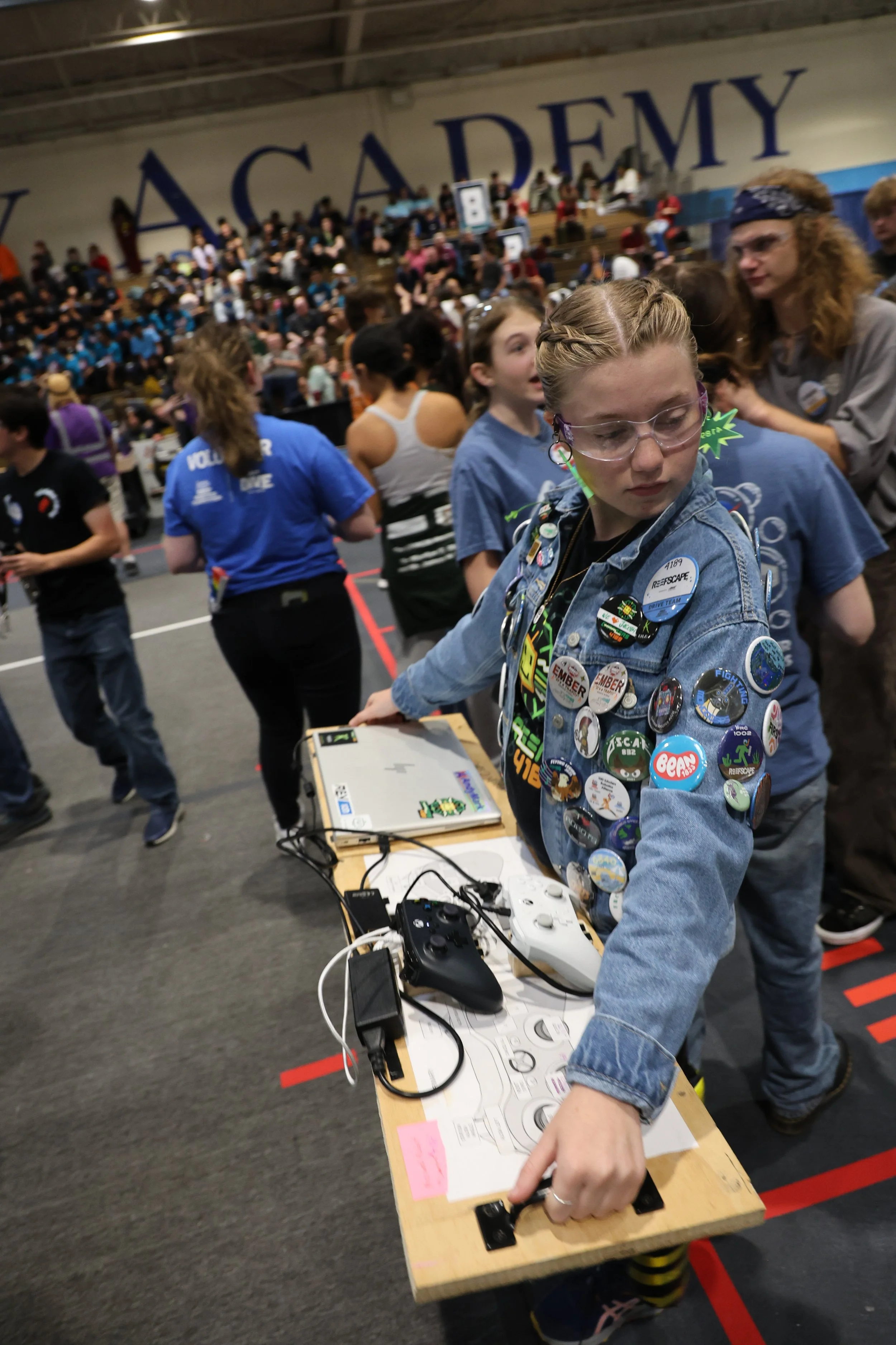 A girl in a denim jacket covered with Girl Scout badges playing a video game at a robotics competition with people and spectators in the background.