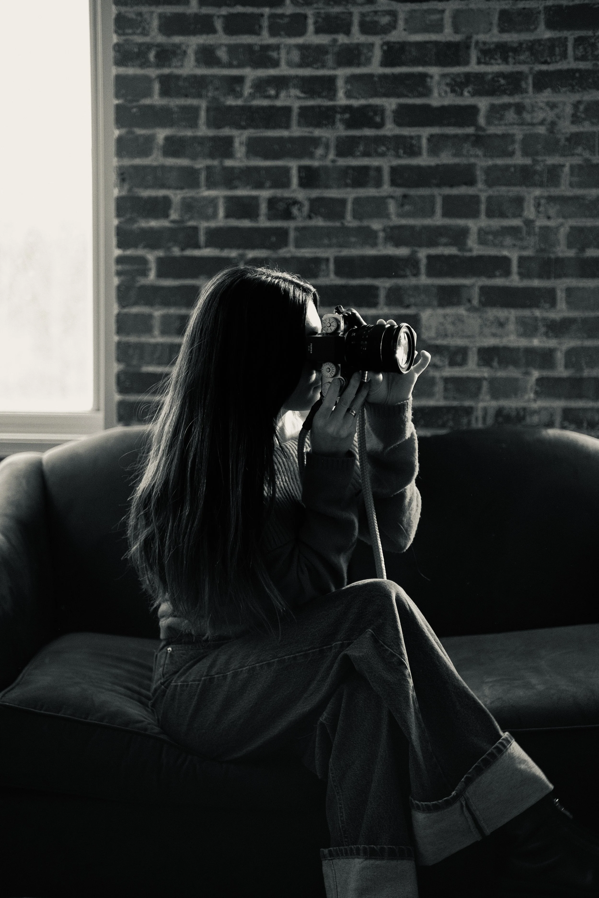 Une femme assise sur un canapé en tissu près d'une fenêtre, tenant un appareil photo et la regardant à travers le viseur, dans une pièce avec un mur en briques.