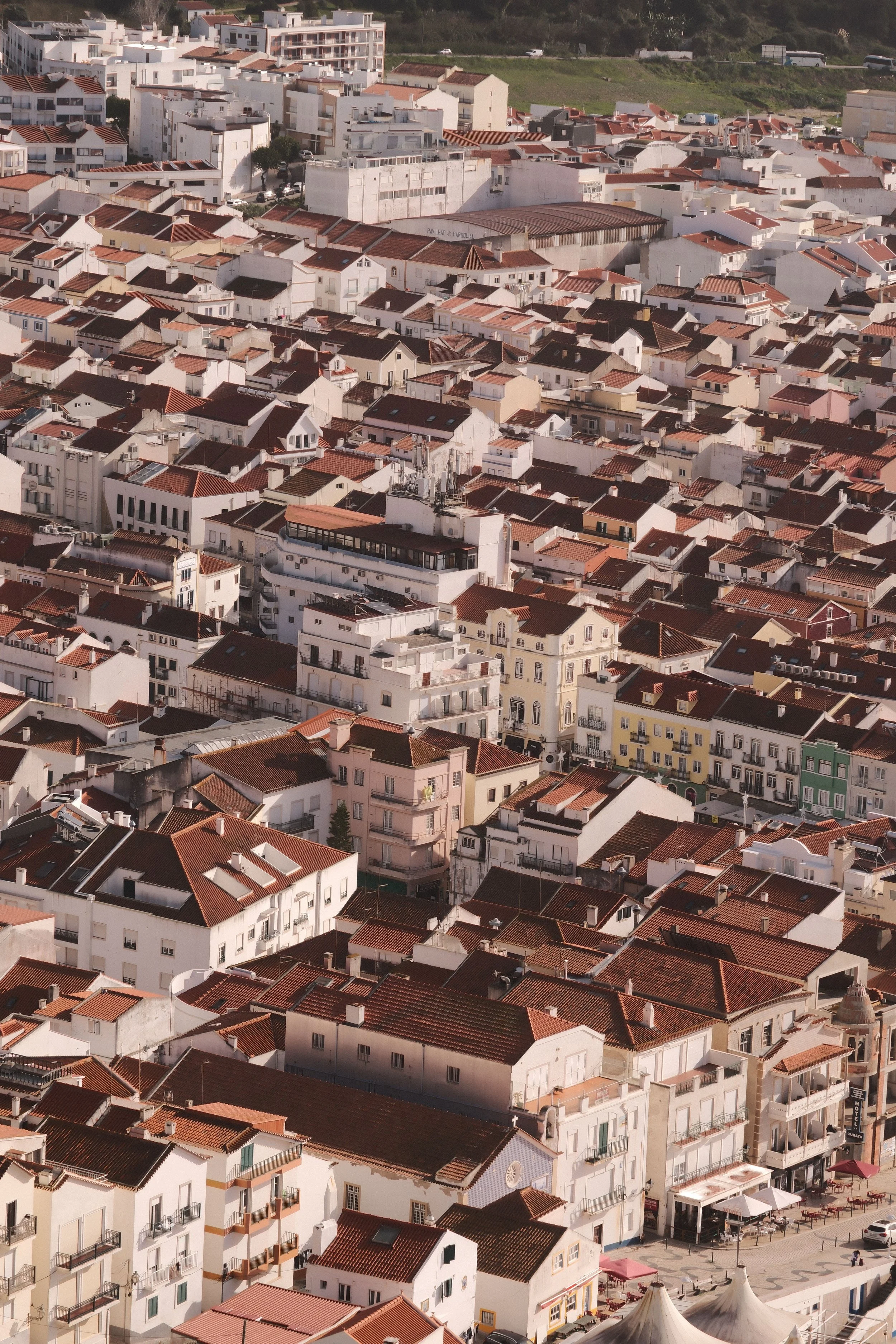 Une vue aérienne d'une ville avec de nombreux bâtiments résidentiels aux toits en tuiles rouges, certains bâtiments plus grands et colorés, avec des rues piétonnes et un espace public en bas de l'image.