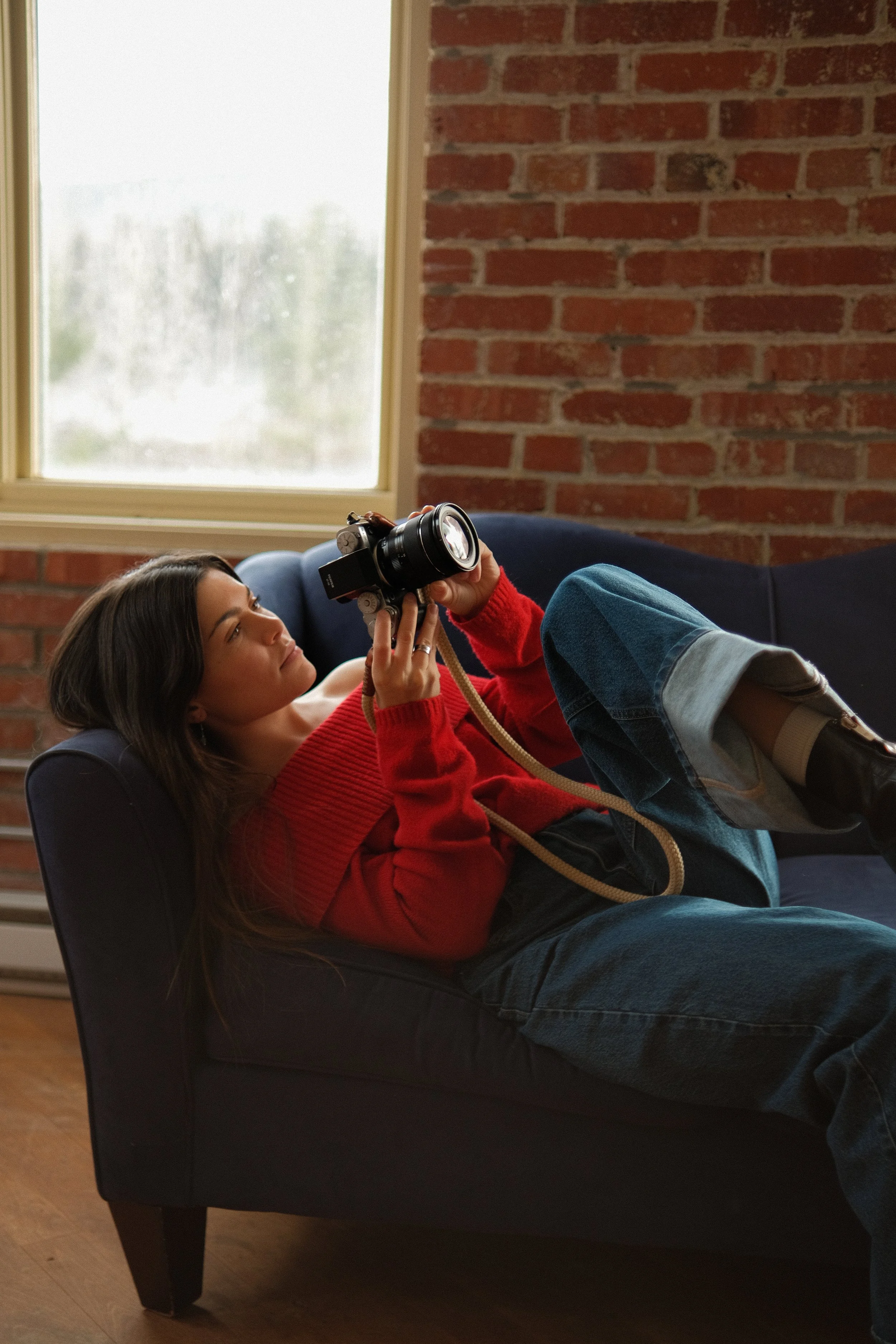 Une jeune femme allongée sur un canapé en train de regarder un appareil photo qu'elle tient.