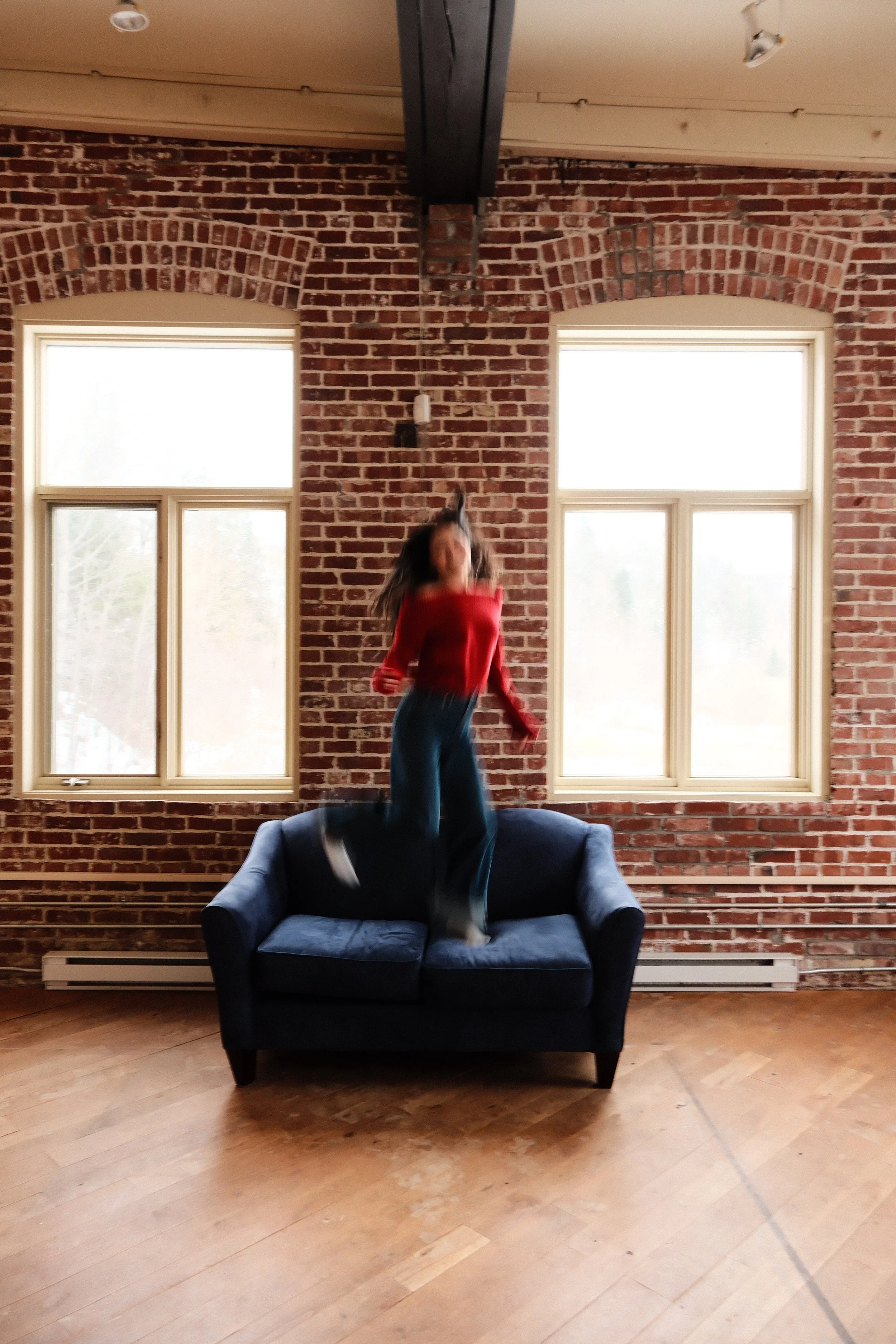 Une femme sautant sur un canapé en velours bleu dans une pièce avec murs en briques et fenêtres avec vue extérieure