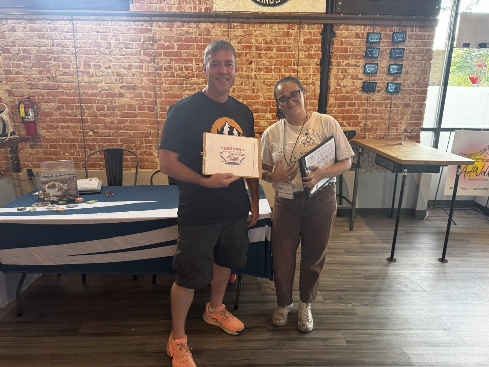 Two smiling people holding CI-133 petitions stand in front of a table at an event in a restaurant.