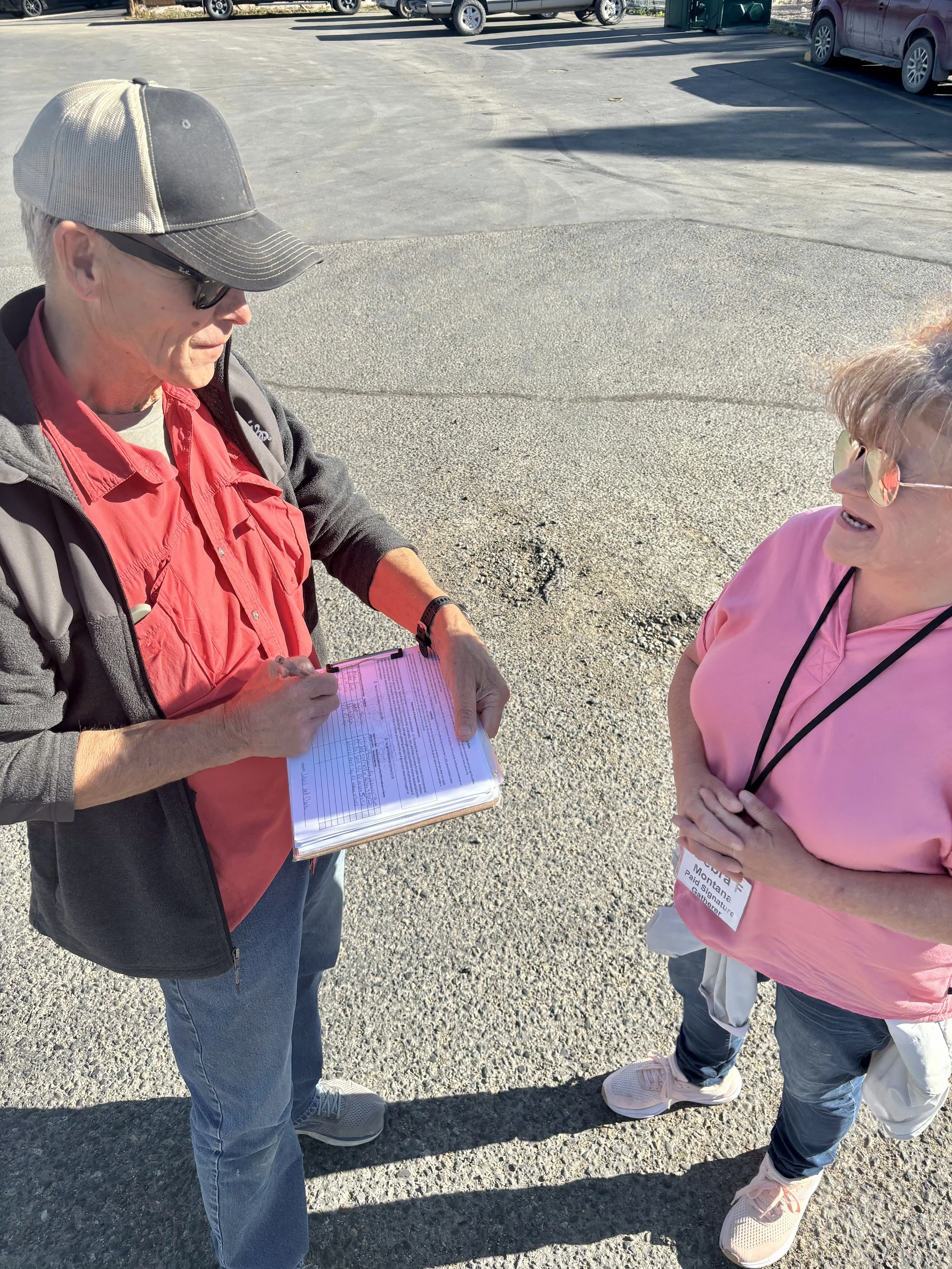 A man wearing a hat and sunglasses talks with a woman wearing a nametag while he signs a CI-133 petition
