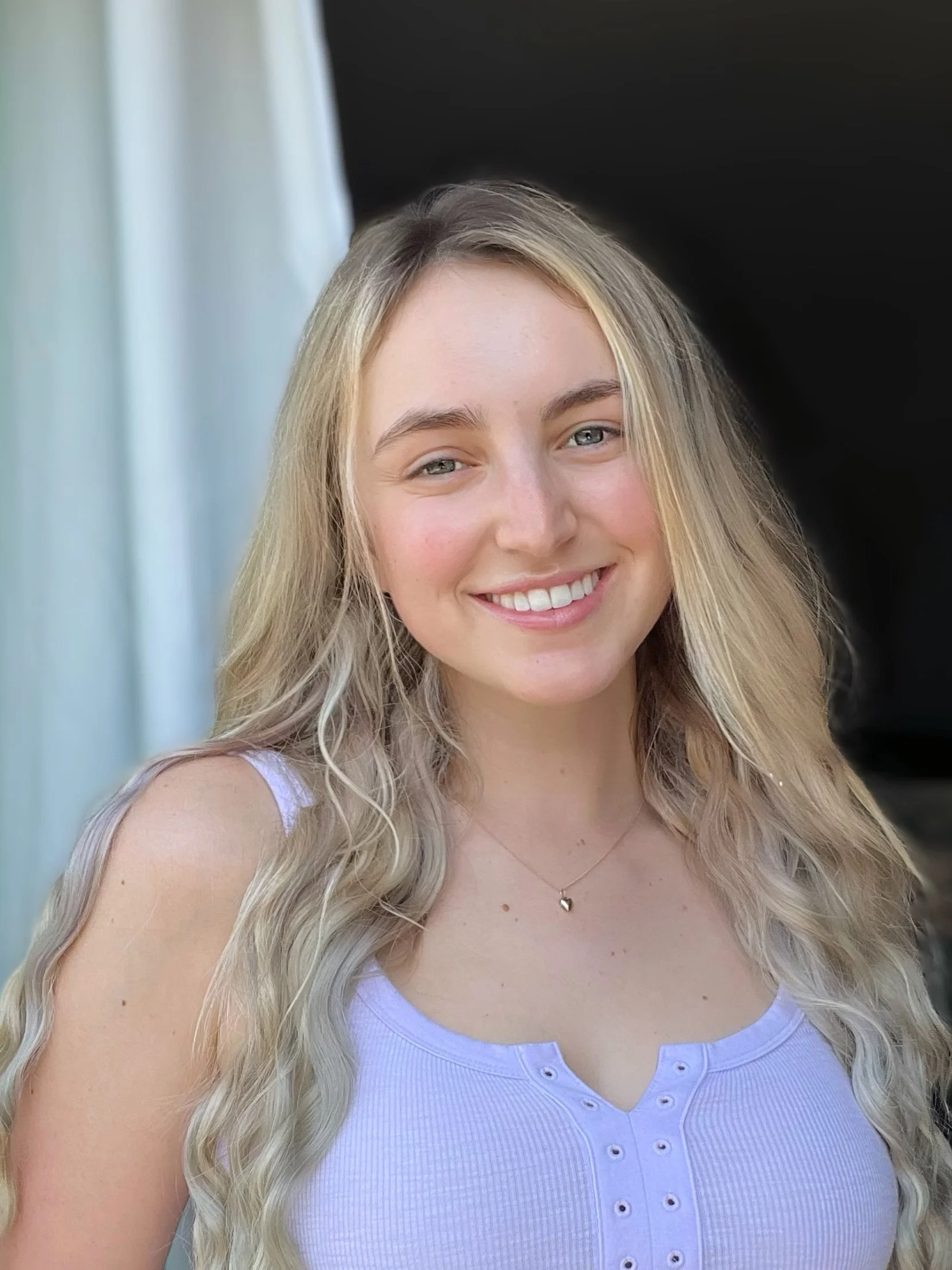 A young woman with long blonde hair, smiling, wearing a white sleeveless top with small buttons, and a delicate heart-shaped necklace.