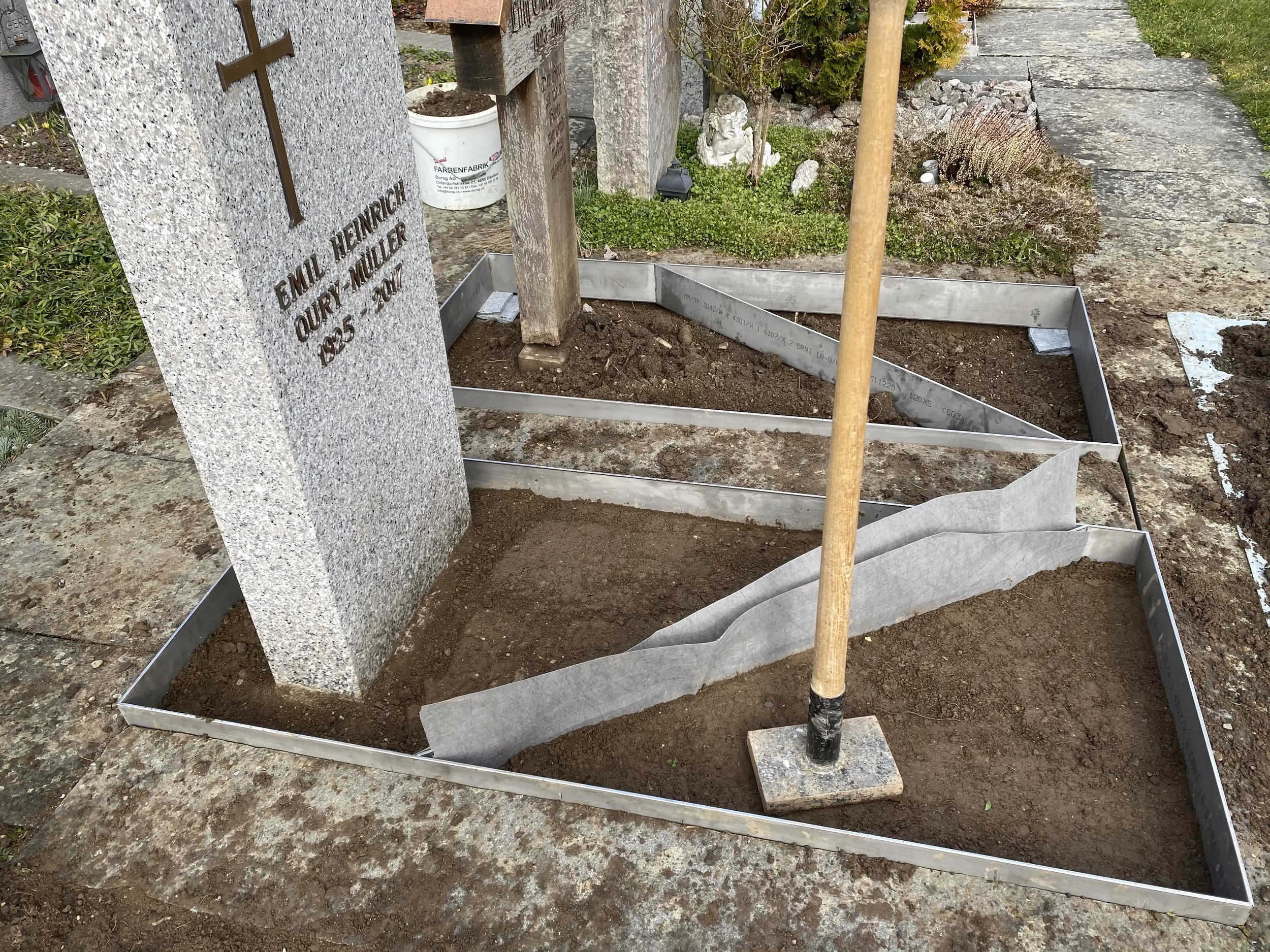 Grabstätte mit einem Grabstein, auf dem steht: Emil Heinrich Outrÿ Müller 1925 - 2001. Der Bereich ist vorbereitet für die Bepflanzung oder Pflege, umgeben von einer Metallrahmenkonstruktion und mit Erde gefüllt.
