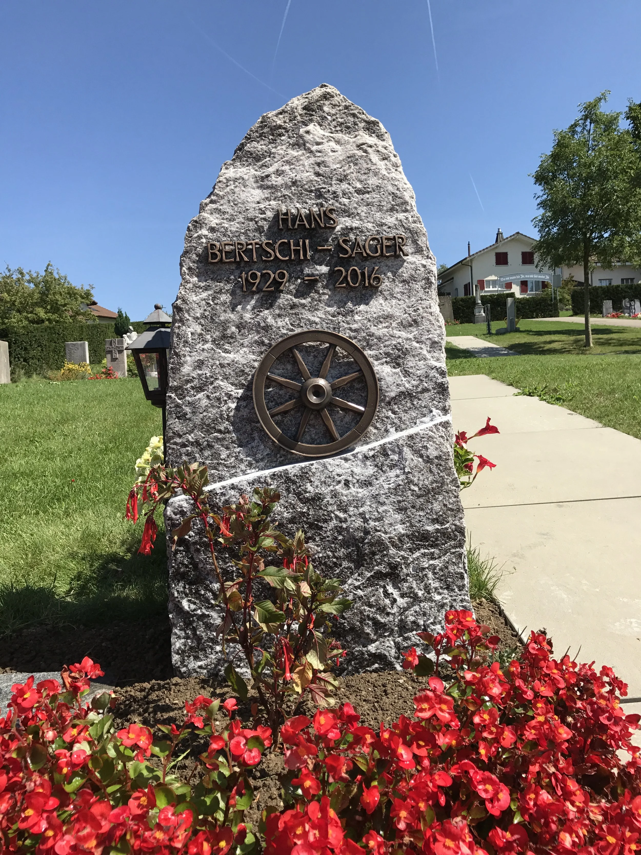 Grabstein mit den Namen Hans Bertschi-Sager, seinen Lebensdaten 1929-2016, und einem Windmühlen-Emblem, umgeben von roten Blumen und auf einer Rasenfläche im Garten.
