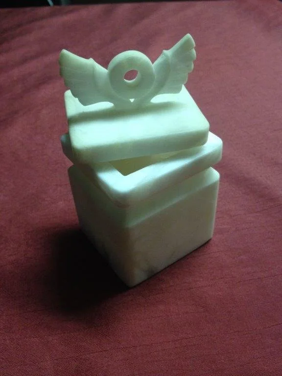 Ein steinernes oder keramisches Behältnis mit einem dekorativen weißen Vogel auf der Deckel, das auf einem braunen Stoff liegt.