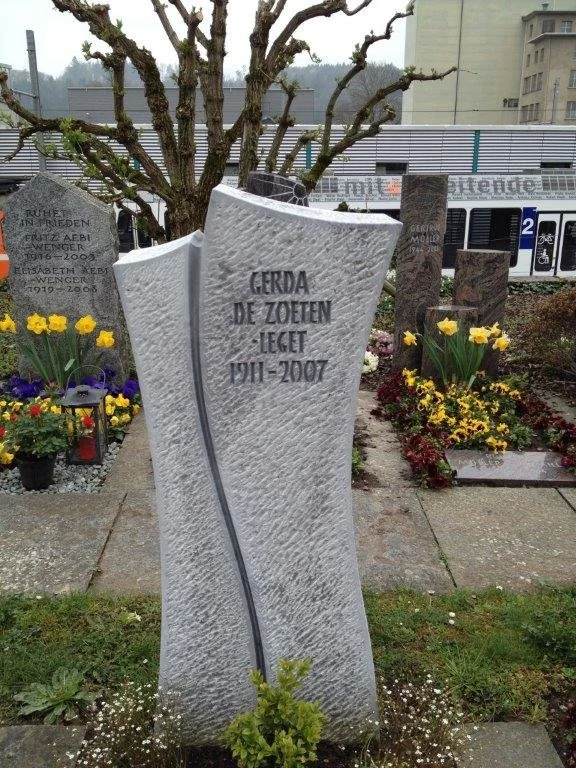 Ein ungewöhnlicher grabstein aus weißem Granit, der schief steht und eine Linie durchquert. Es sind Blumen und ein Baum im Hintergrund zu sehen.