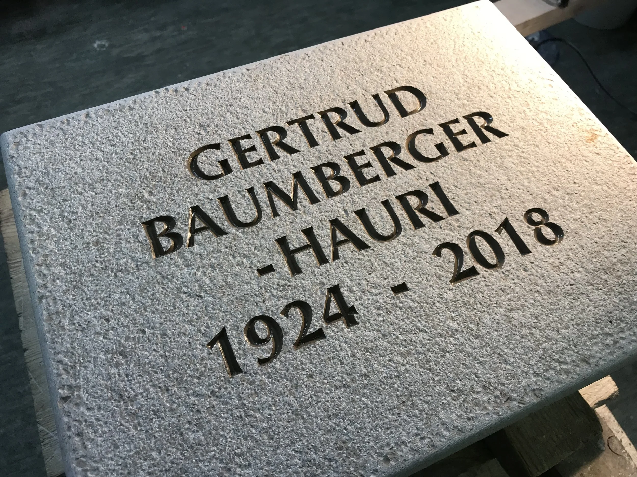 Gedenktafel mit dem Text: GERTRUD BAUMBERGER-HAURI 1924 - 2018 auf einer steinernen Oberfläche.