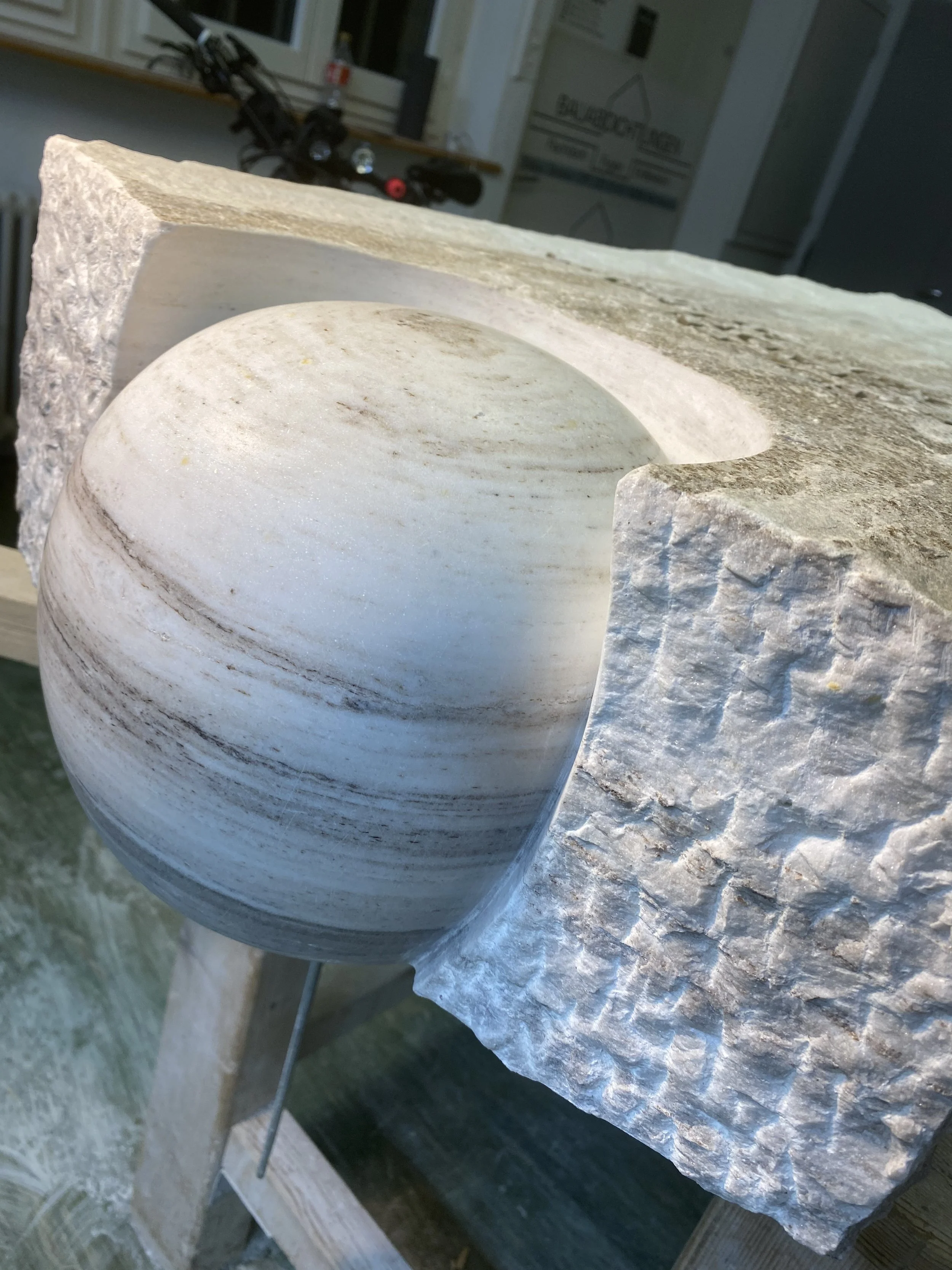 Künstlerische Skulptur aus Sandstein mit einer glatt geschliffenen Oberfläche, auf einem Holzgestell präsentiert.