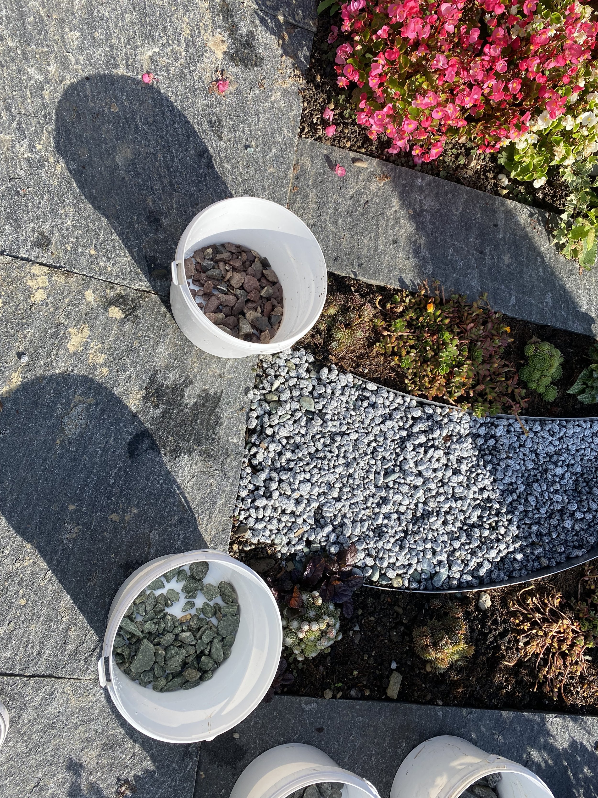 Zwei weiße Eimer mit Steinen, eine Gartengestaltung mit Erde, bunten Pflanzen und Kieselsteinen, umgeben von großen Steinen und einer Blumenbühne mit pinken und weißen Blumen.