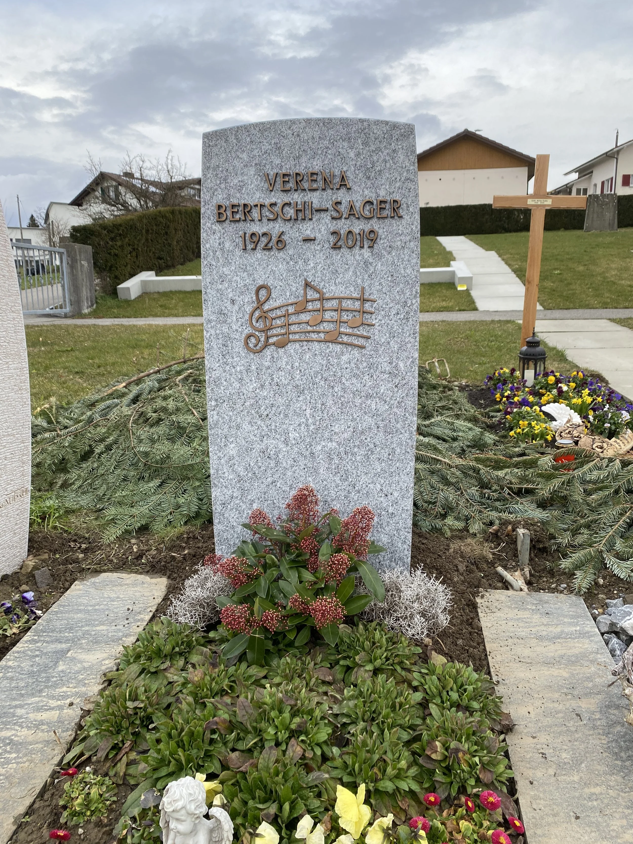 Grabstein mit dem Namen Verena Bertschi-Sager, Geburtsjahr 1926 und Sterbedatum 2019, mit musikalischem Notenschlüssel und Noten, Blumen und einem kleinen Engelchen davor, in einem Garten.