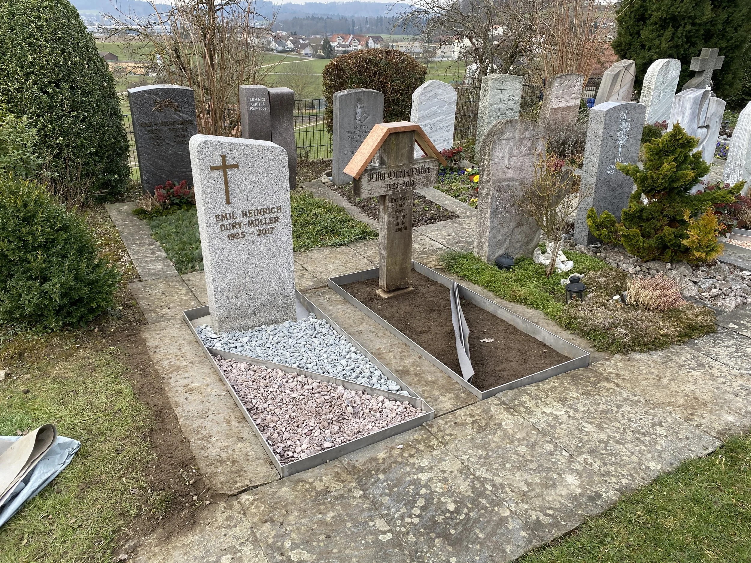 Grabstätte mit zwei Grabsteinen, eine davon mit Kreuz und Namen Emil Heinrich Oury-Müller, daneben ein provisorisches Holzkreuz für Lily Oury-Müller, umgeben von Blumen und Bäumen auf einem Friedhof, mit einer ländlichen Landschaft im Hintergrund.