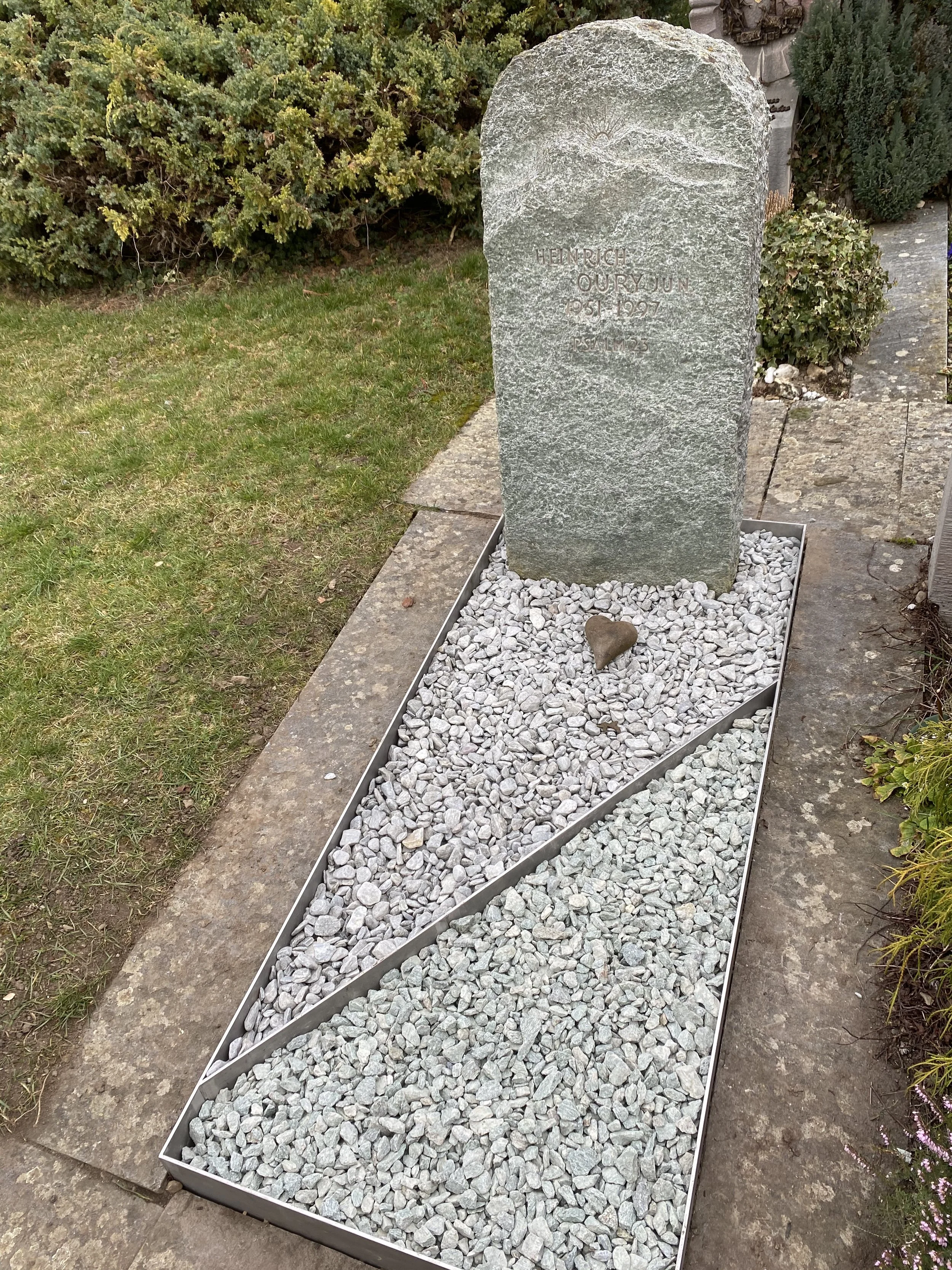 Granitgrabstein mit Beschriftung für Heinrich Ouryun auf einem bepflanzten Friedhof, umgeben von grau-weißen Kieselsteinen in einer metalldosenförmigen Begrenzung.