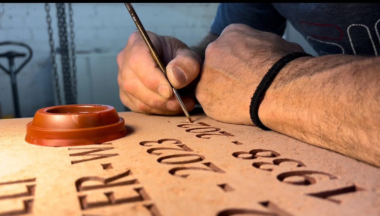 Person schreibt mit einem Pinsel auf einem Holzbrett mit eingravierten Buchstaben.