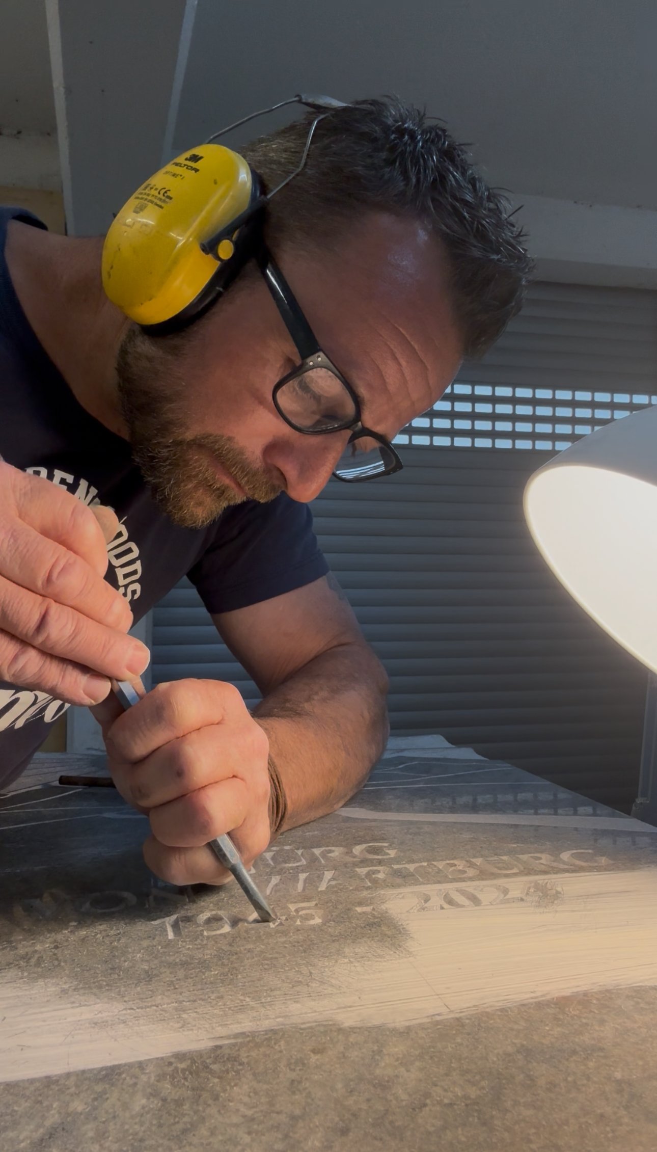 Mann arbeitet mit einem Stift auf einem Holzbrett, trägt Schutzbrille und Gehörschutz.