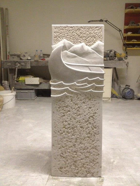 Eine Skulptur in Form eines stilisierten Schiffs mit Wellen, die aus hellem Stein herausgemeißelt ist, in einer Werkstatt.