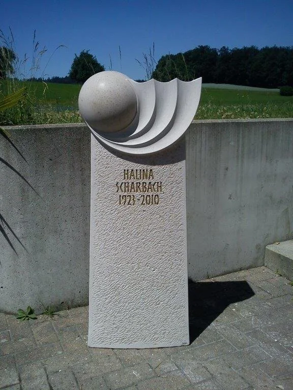 Gedenkstein mit einer Skulptur aus einem halbrunden und kugelförmigen Element sowie hervorschauenden Schichten, mit der Inschrift "Hauna Scharbach 1922-2010".