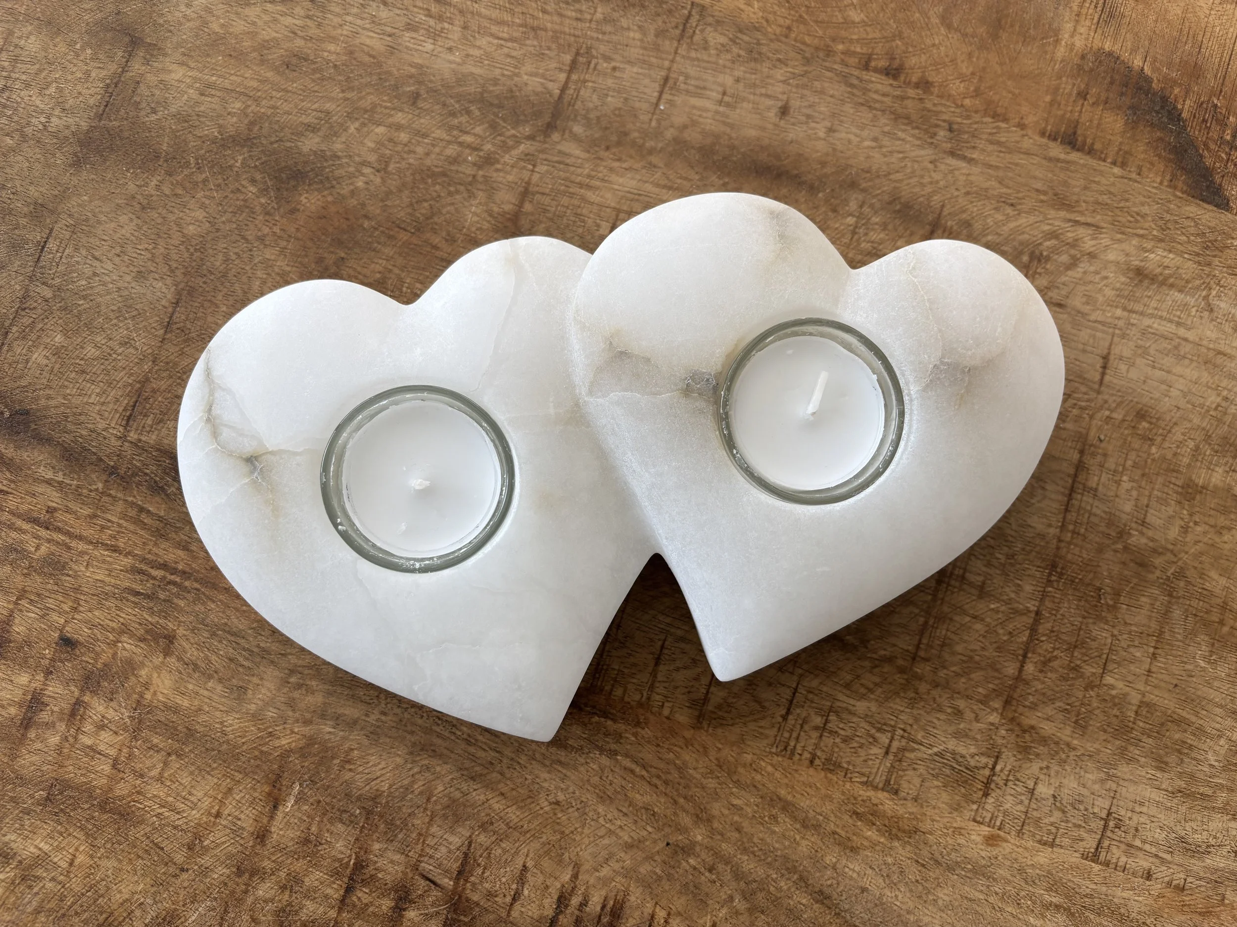 Zwei Kerzen in kleinen Gläsern, die auf einem weißem, herzförmigem Kerzenhalter aus Marmor sitzen, auf einem Holztisch.