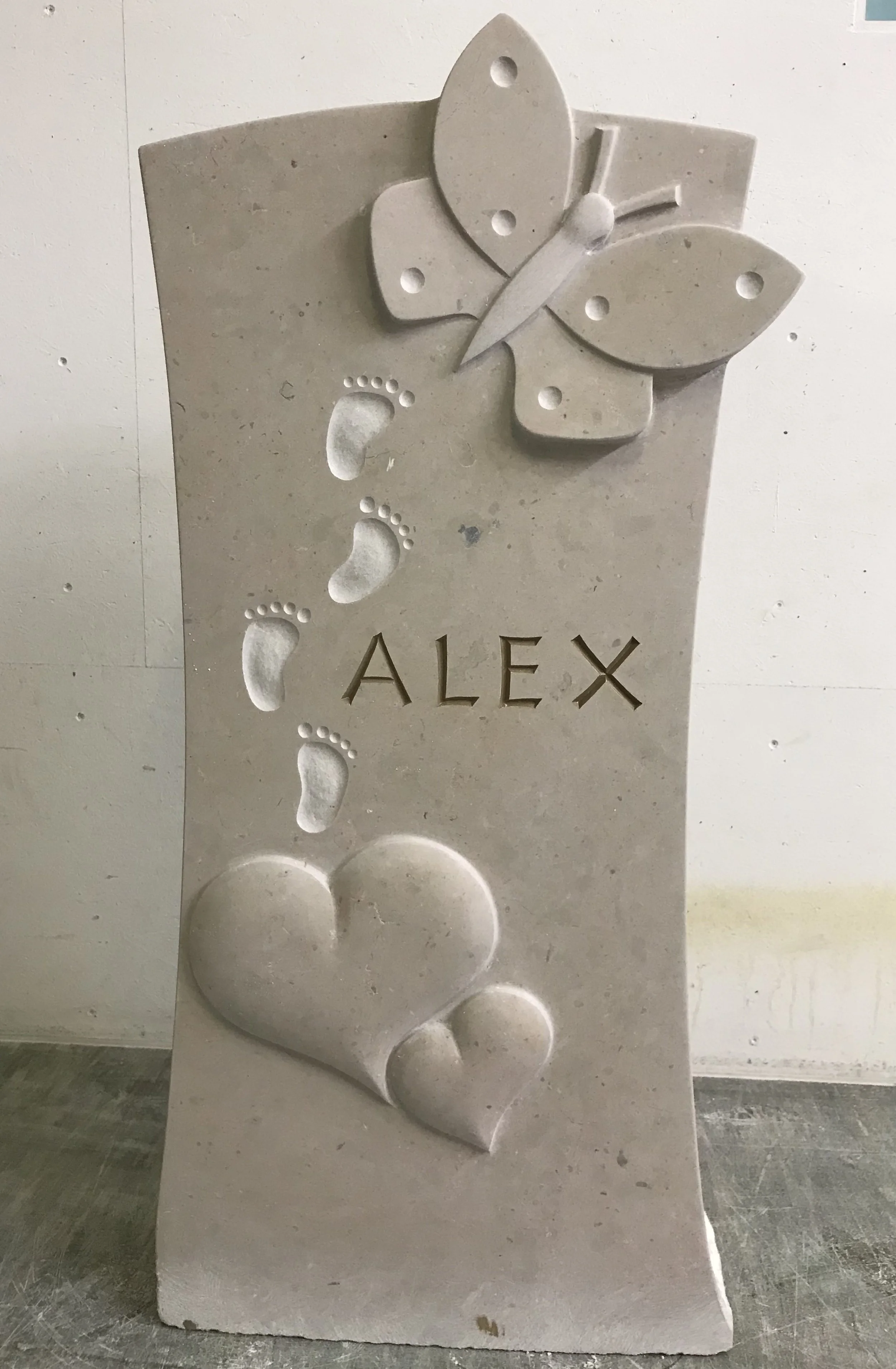 Eine Grabsteinskulptur aus hellem Stein mit einem Schmetterling, Fußspuren, dem Namen 'ALEX' und zwei Herzen.