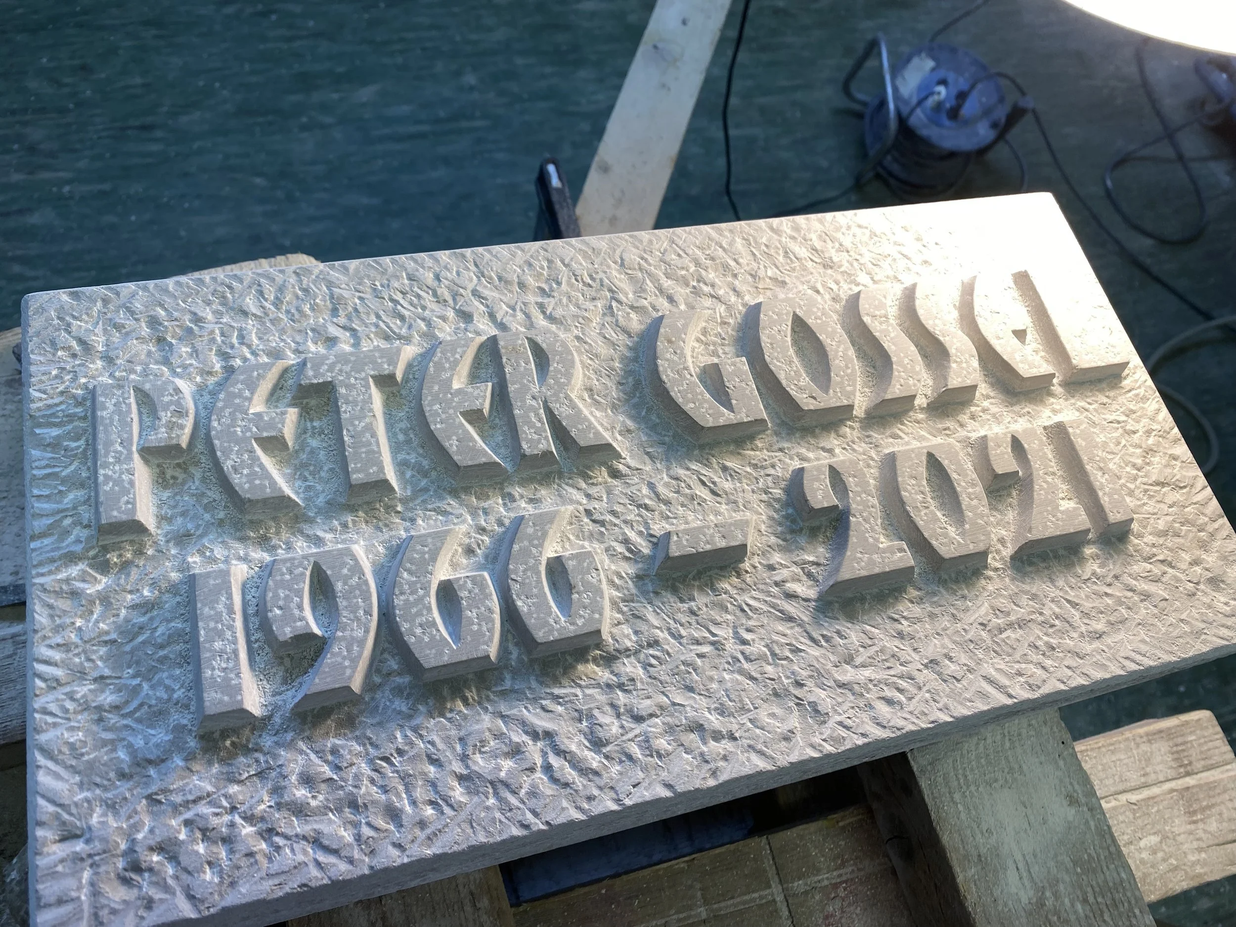 Holzblock mit in 3D geschnitzten Worten und Zahlen, die 'PETER GOSSER 1960-2021' anzeigen.