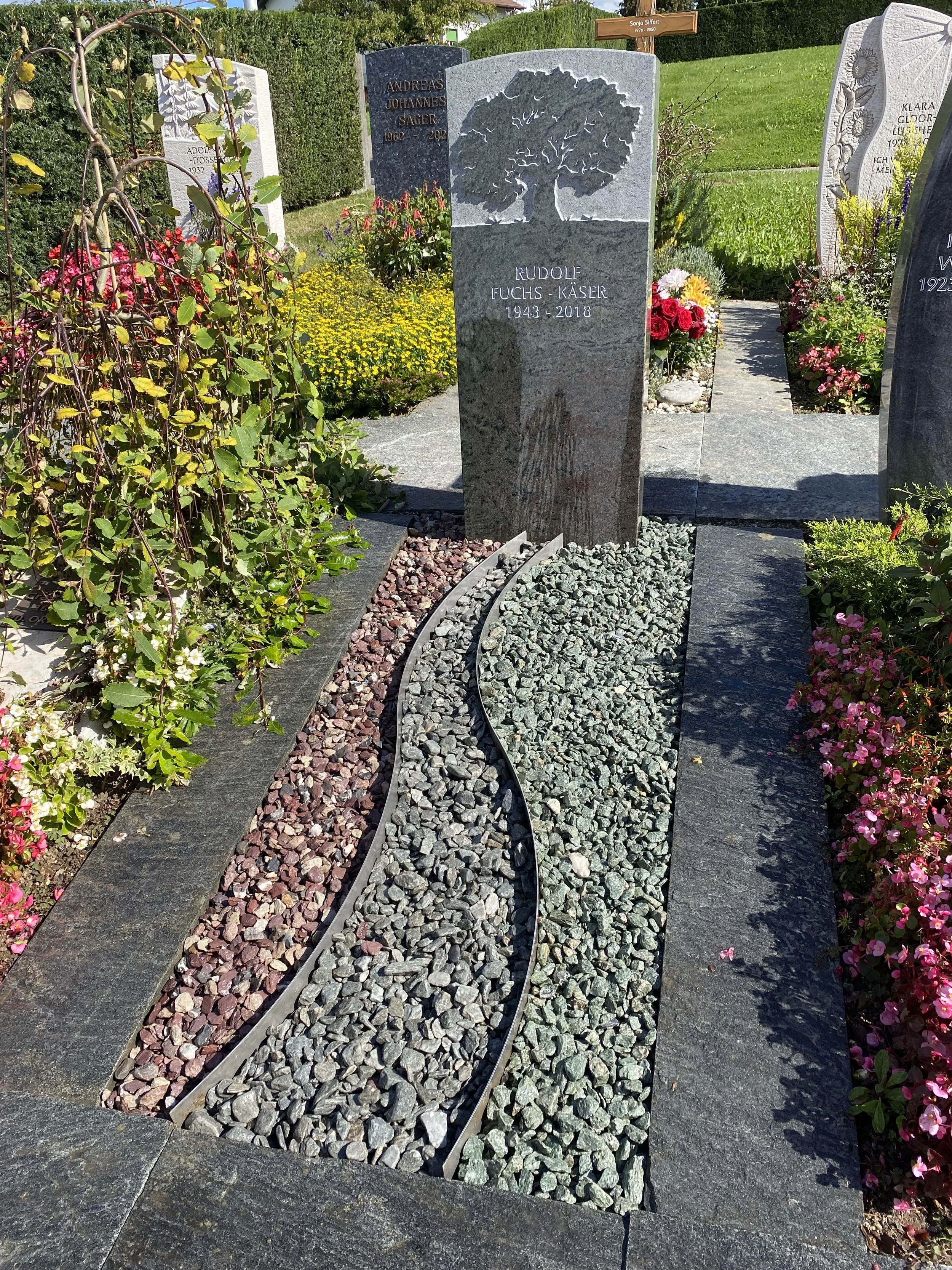 Grabstein in einem gepflegten Friedhof mit Blumen und dekorativen Steinen, inschriftlich für Rudolf Fuchs-Käser, geboren 1943 und verstorben 2018, mit Baum-Relief oben.