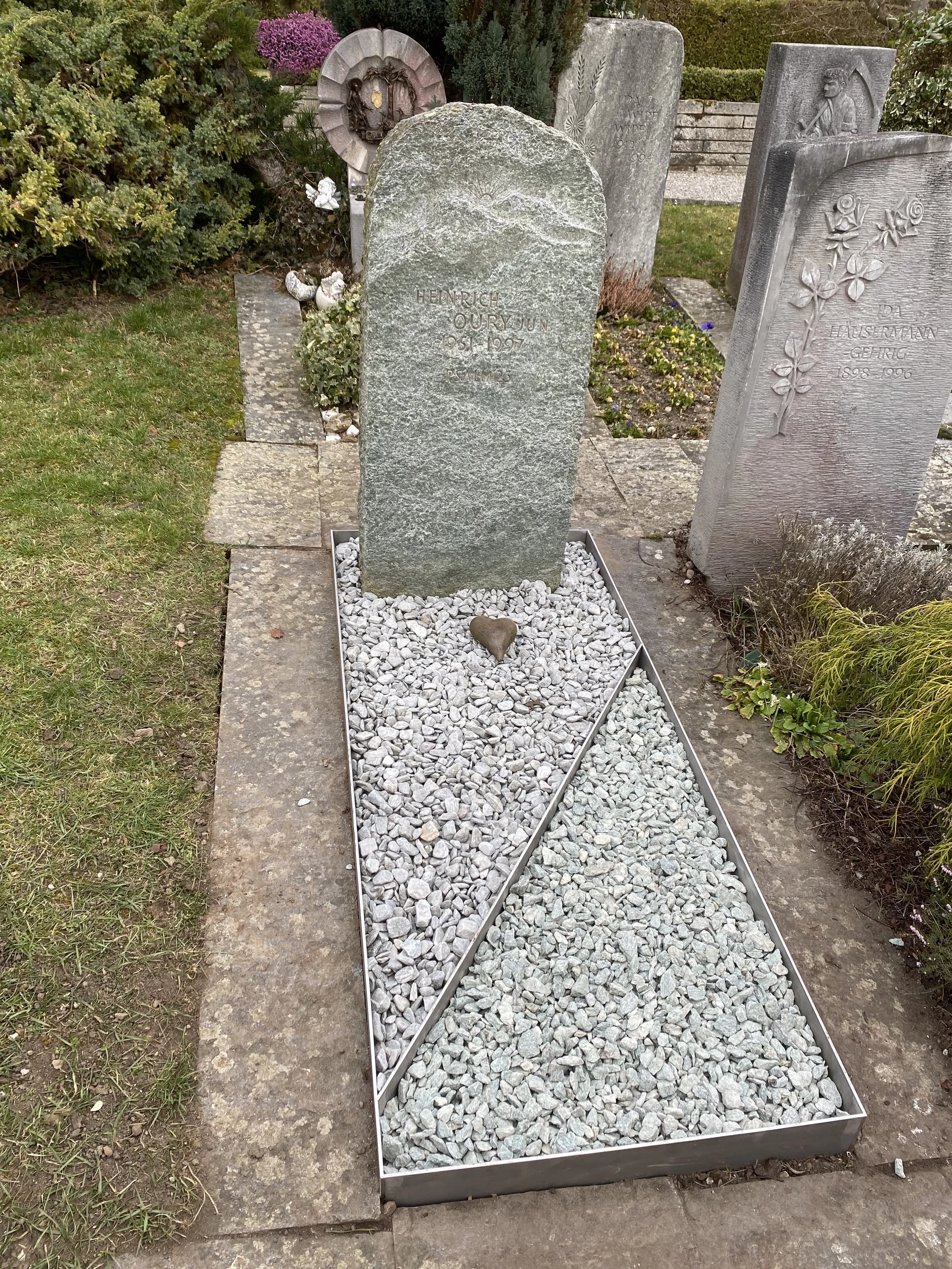 Ein Grabschfeld mit Steinen und einem Grabstein, auf dem steht: 'HEINRICH OURY JUN 05.11.1907'. Das Grab ist mit weißen Kieseln und einem kleinen herzförmigen Stein bedeckt.