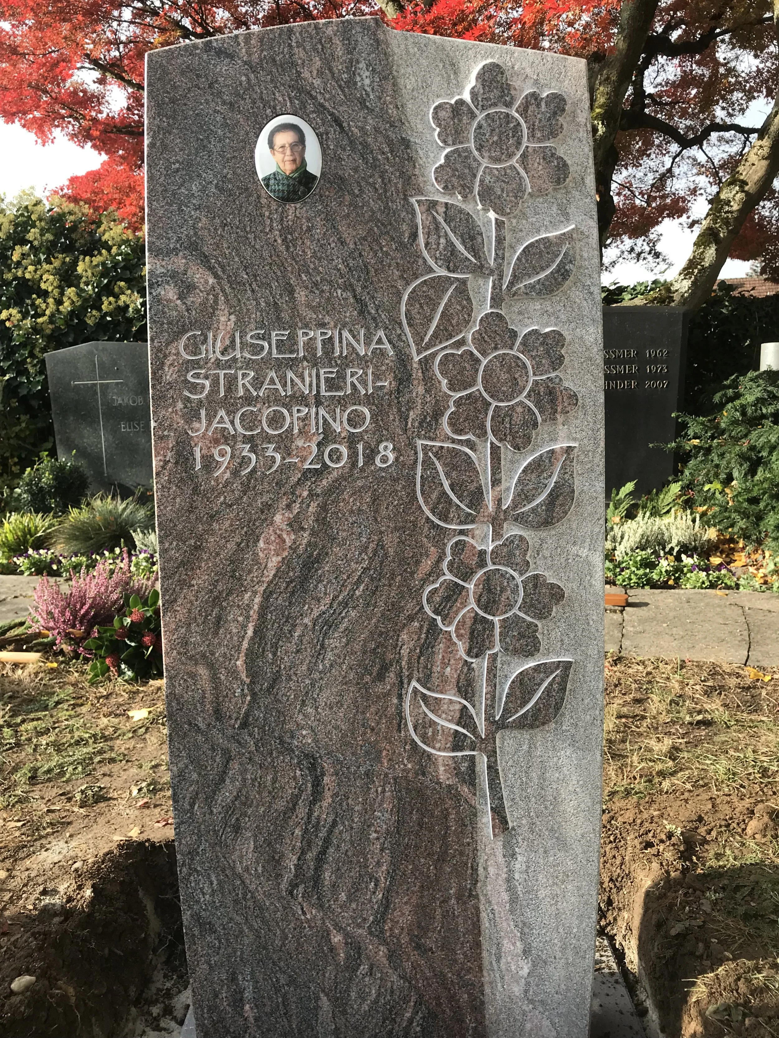 Grabstein mit einem Porträtfoto einer älteren Frau, eingravierten Blumen und Blättern, mit dem Namen GUSEPPINA STRANERI-JACOPINO sowie den Jahren 1955-2018, im Friedhofsbereich mit Blumen und Bäumen im Hintergrund.