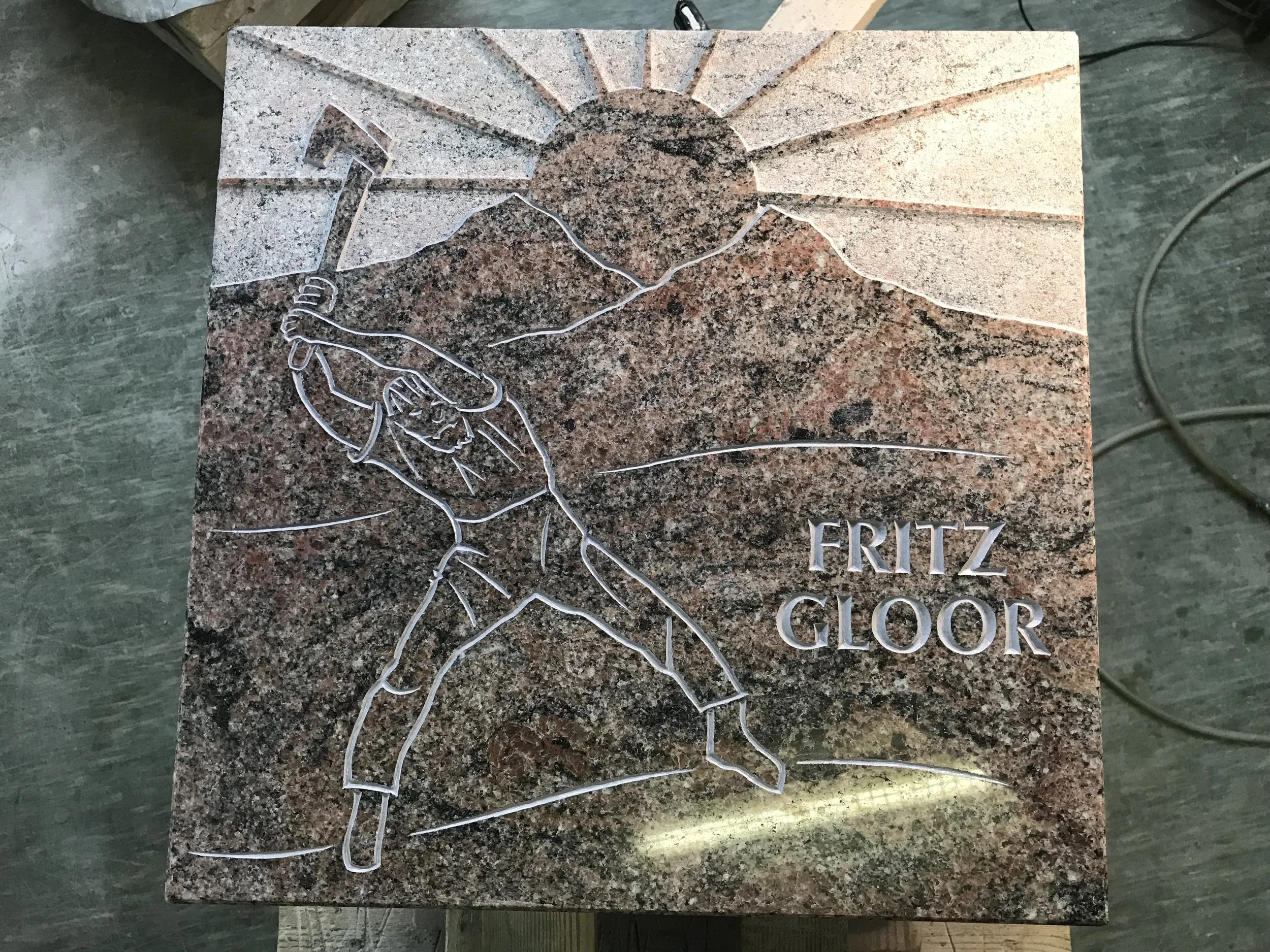 Stein mit eingravierter Darstellung eines Mannes, der einen Hammer hebt, und der Sonne im Hintergrund. Der Name 'Fritz Gloor' ist eingraviert.