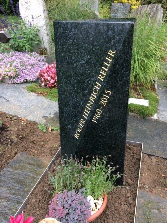 Ein schwarzer Grabstein in einem Garten mit dem Namen "Roger Heinrich Reiffer", den Jahren 1960 bis 2015, besteht aus Marmor oder Granit, umgeben von Blumen und Pflanzen.