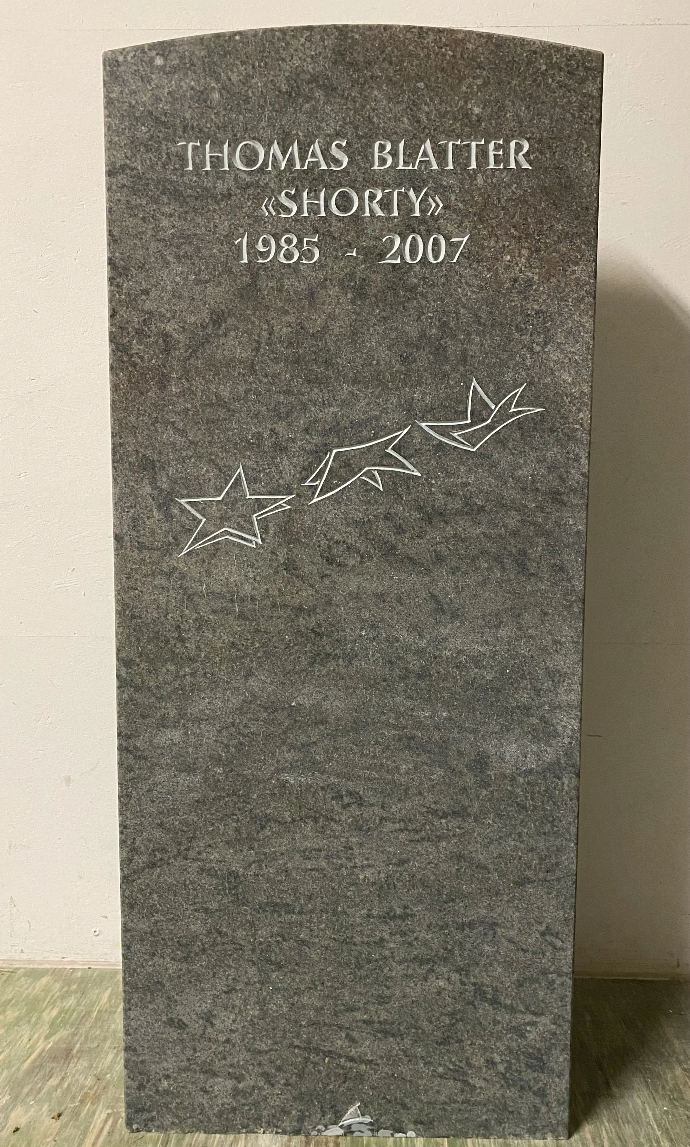 Grabstein mit eingraviertem Namen Thomas Blatter, Spitzname 'Shorty', Geburtsjahr 1985 und Sterbejahr 2007, mit Dekoration von drei Sternen und zwei Vögeln aus Linienzeichnung.