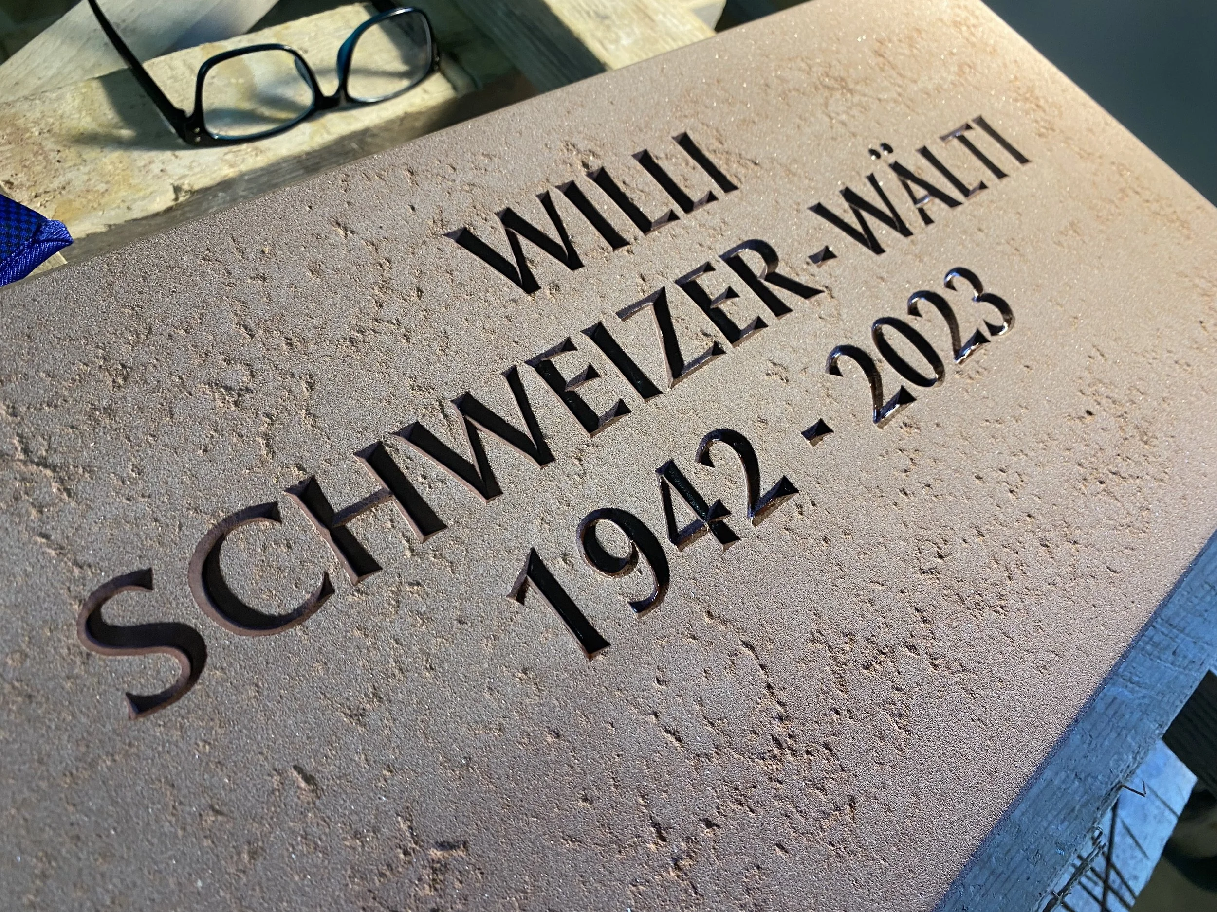 Gedenktafel mit eingraviertem Text 'SCHEWENEGINI WALL' und den Jahren 1942-2023, auf einem Sandstein-Relief, dazu ein Paar Brillengläser, ein blauer Stoff und eine Holzoberfläche.