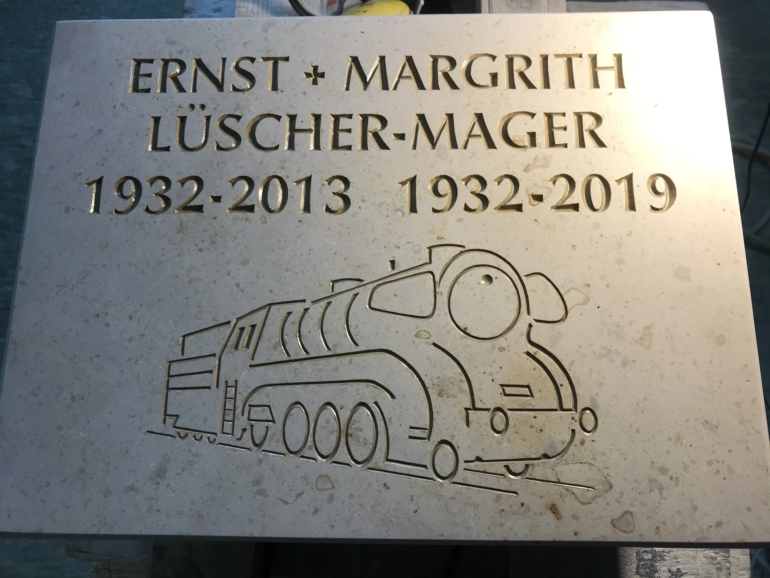 Gedenktafel für Ernst und Margrith Lüscher-Mager mit den Jahren 1932-2013 und 1932-2019, sowie einer stilisierten Lokomotive.