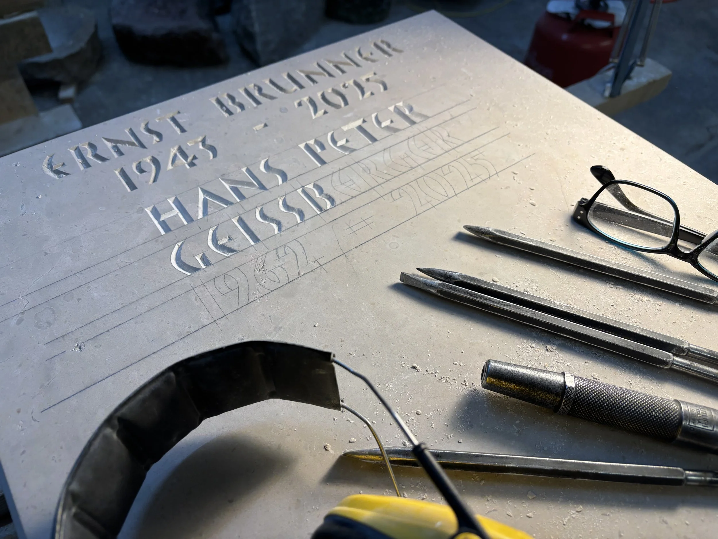 Fräsen eines steinernen Gedenksteins mit gravierten Texten und Skizzen, Werkzeugen und Brille liegen darauf.