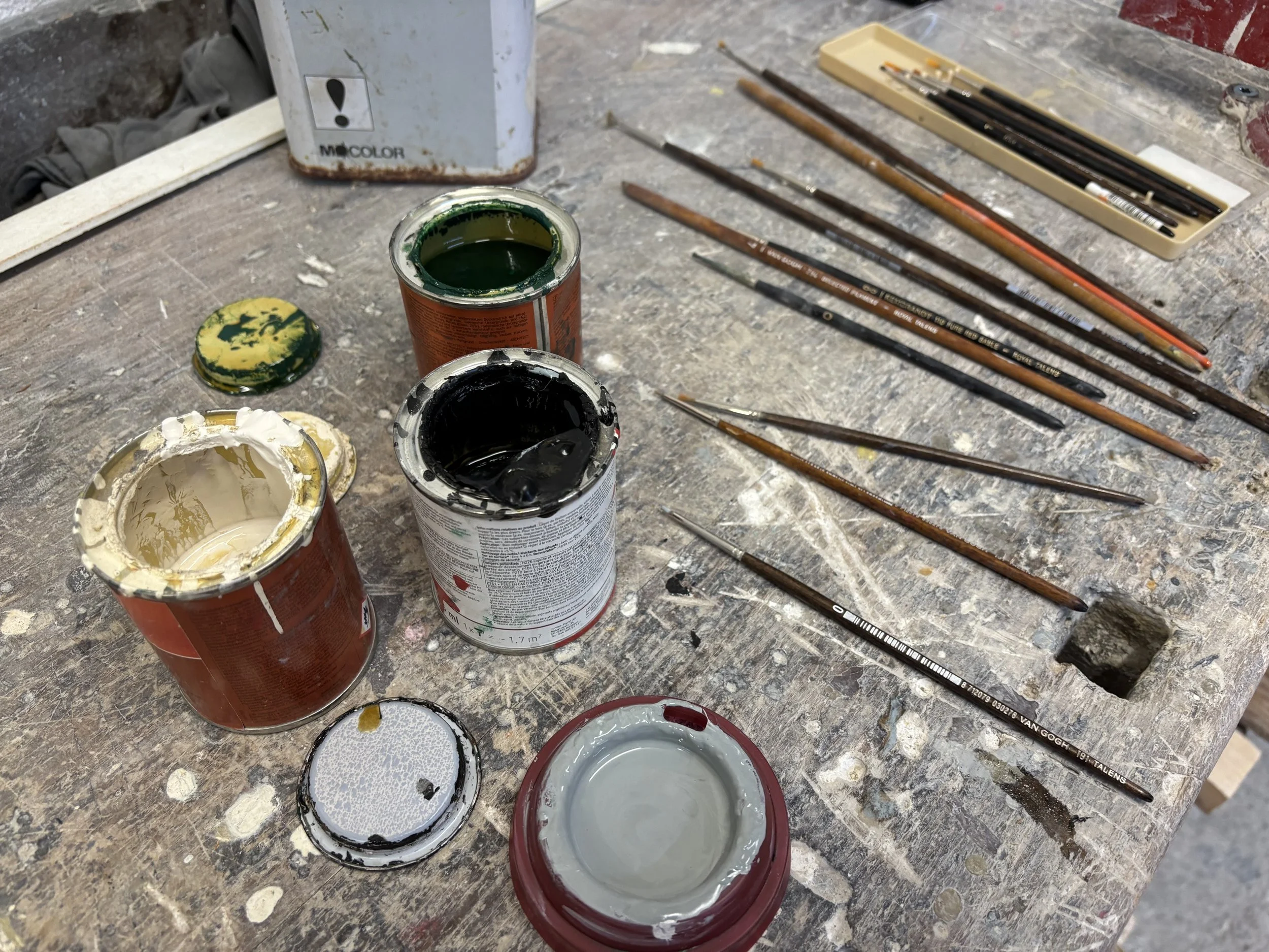 Kunstzubehör auf einem alten, verschmutzten Tisch, inklusive Farben in Dosen, Pinsel und ein Farbpalette in einer Werkstatt.