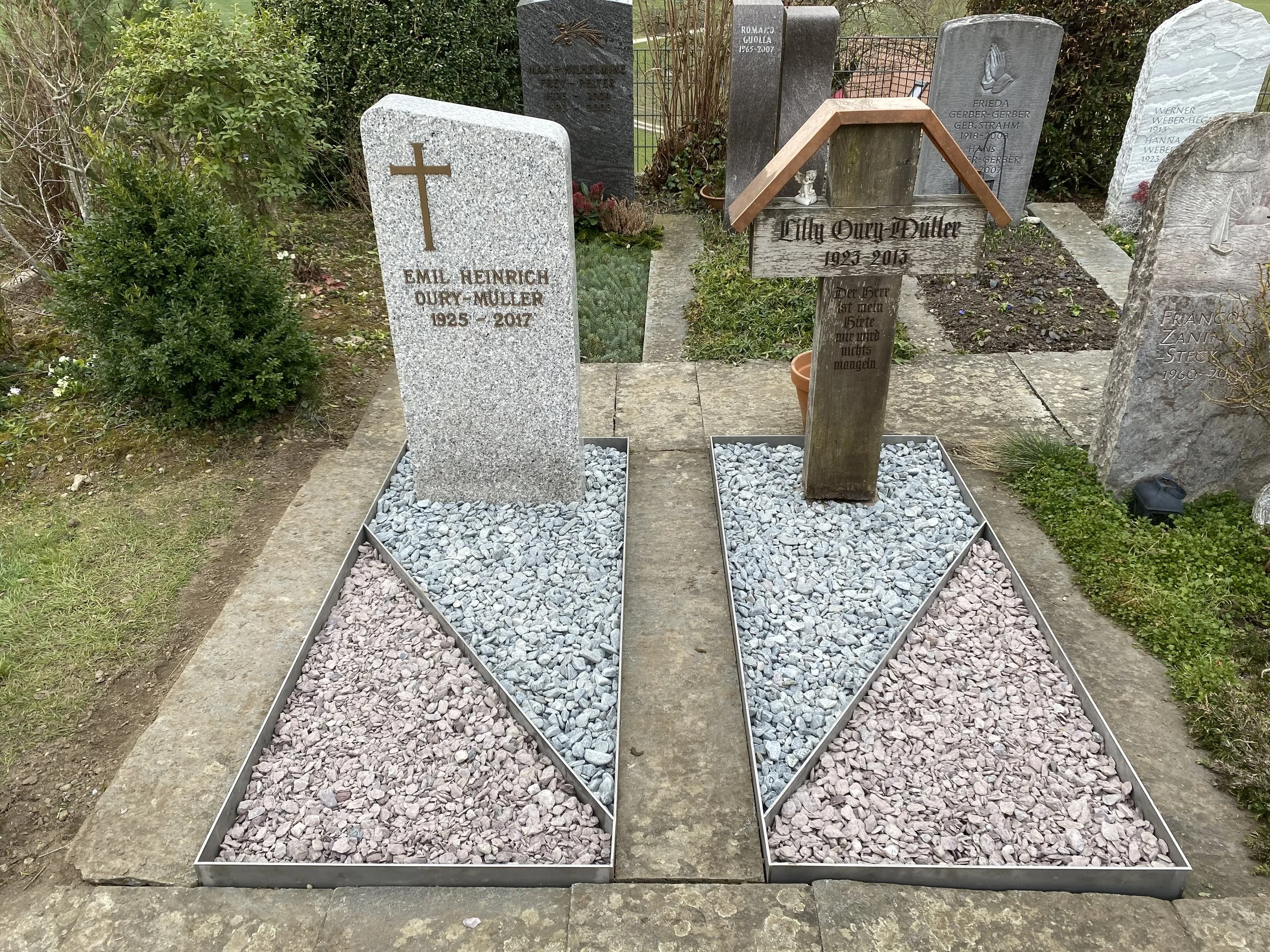 Zwei Gräber mit Grabsteinen, eines aus Granit mit Kreuz, das andere aus Holz mit einem Dach, auf einem Friedhof.