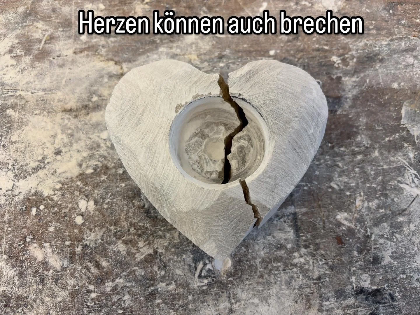Ein zerbrochenes Herz aus Holz mit einer Kerze in der Mitte und einer aufgedruckten deutschen Textüberschrift.
